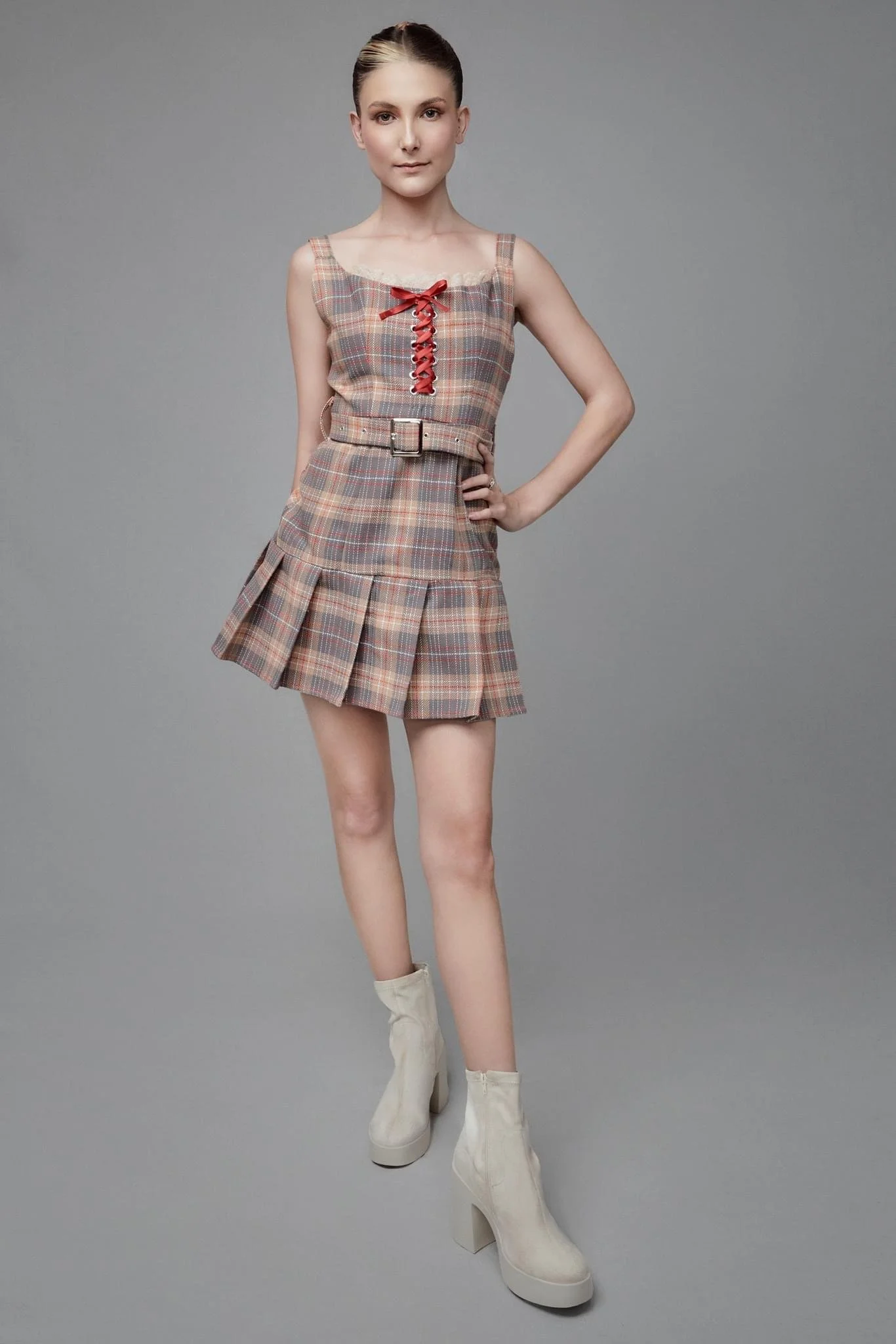 A young woman wearing a plaid sleeveless dress with a red lace-up detail on the chest, a belt at the waist, and a pleated skirt, paired with white ankle boots, standing against a plain gray background.