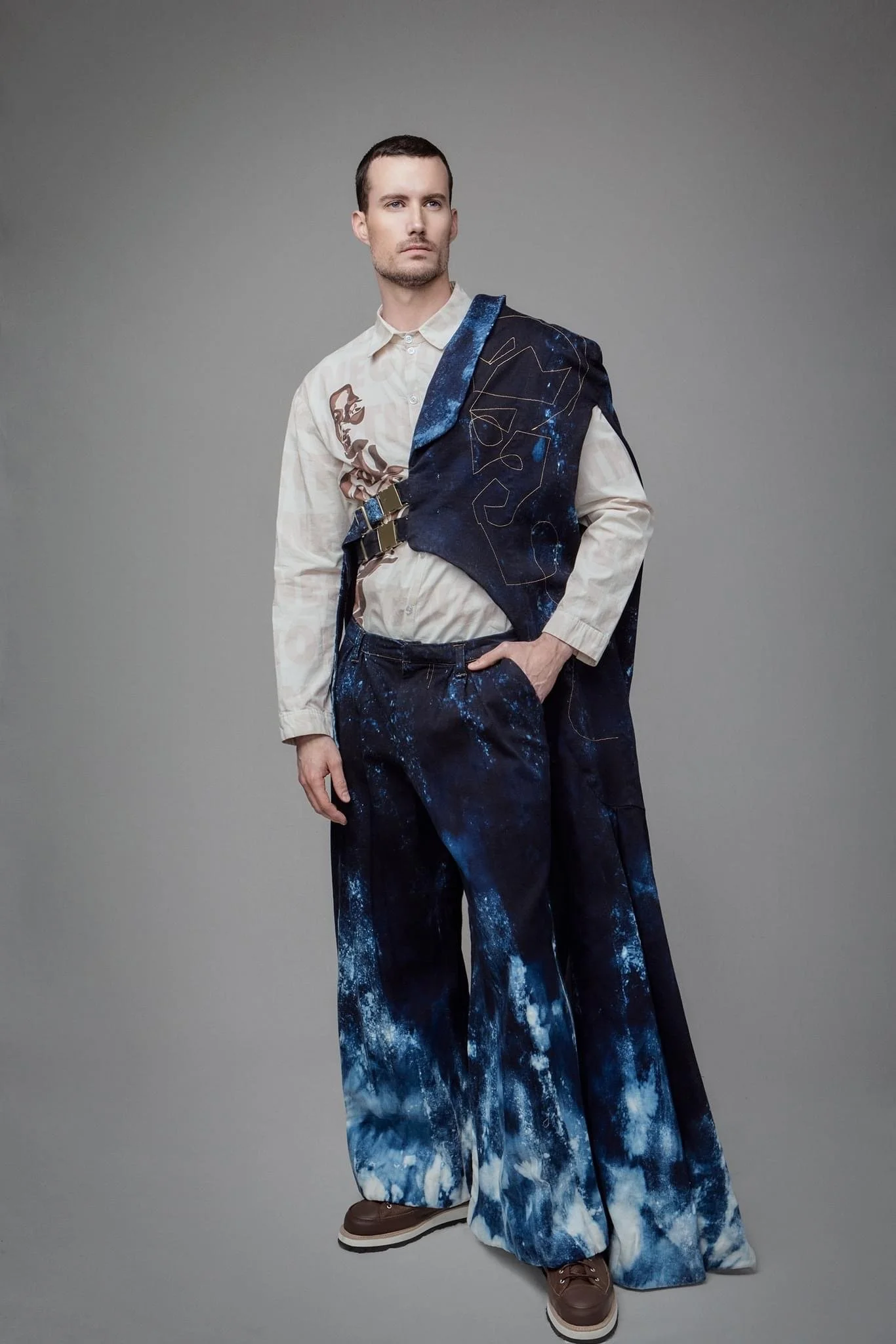 A man wearing a white button-up shirt and loose, wide-leg pants with a blue and white tie-dye pattern, standing against a gray background.