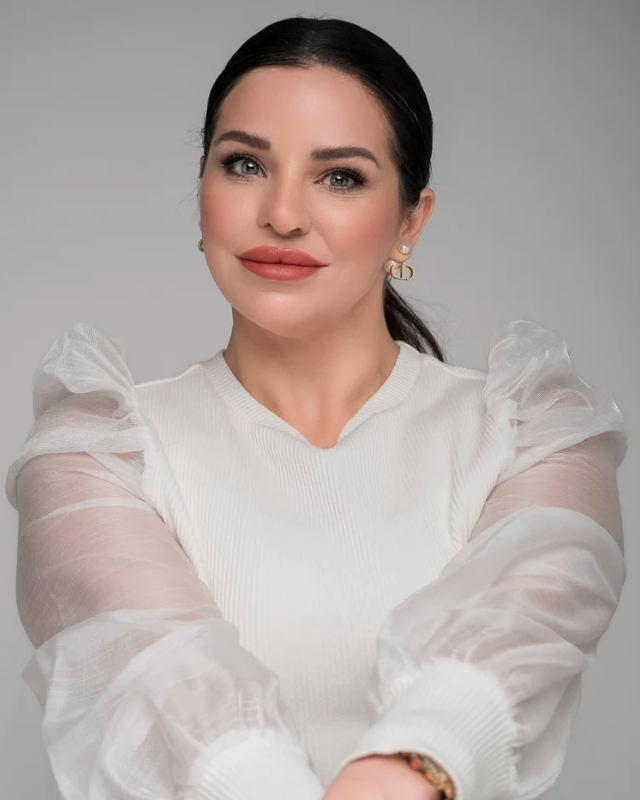 A woman with dark hair pulled back, wearing makeup and gold earrings, dressed in a white top with sheer puffed sleeves, smiling slightly against a gray background.