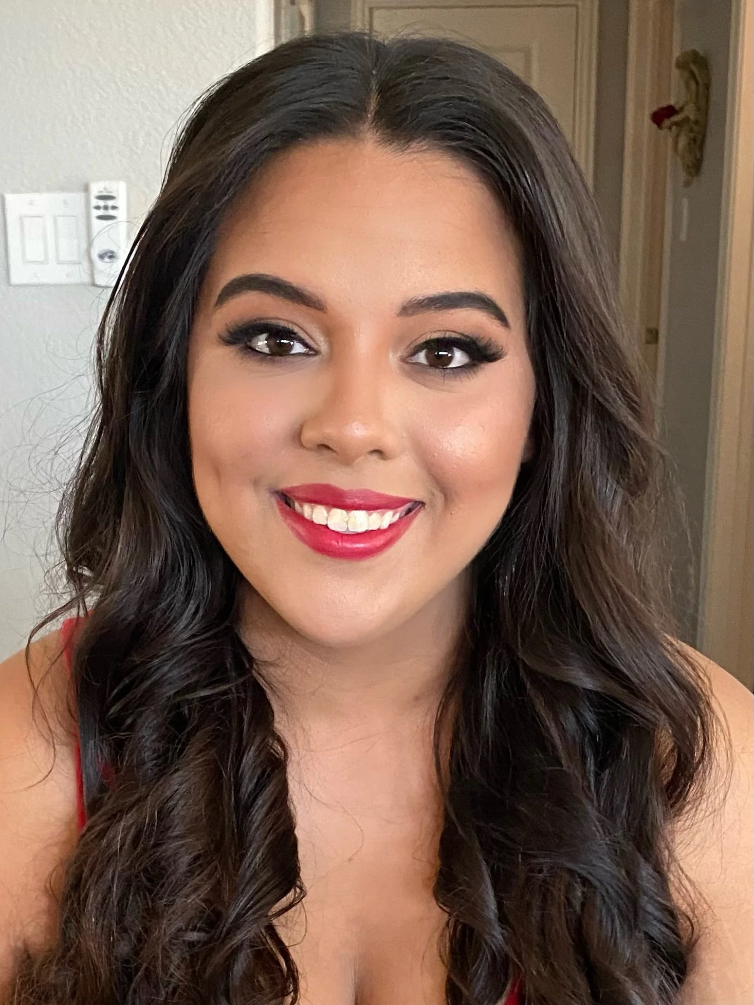 A smiling woman with long dark wavy hair, makeup, and a red top, indoors with a white wall and door in the background.