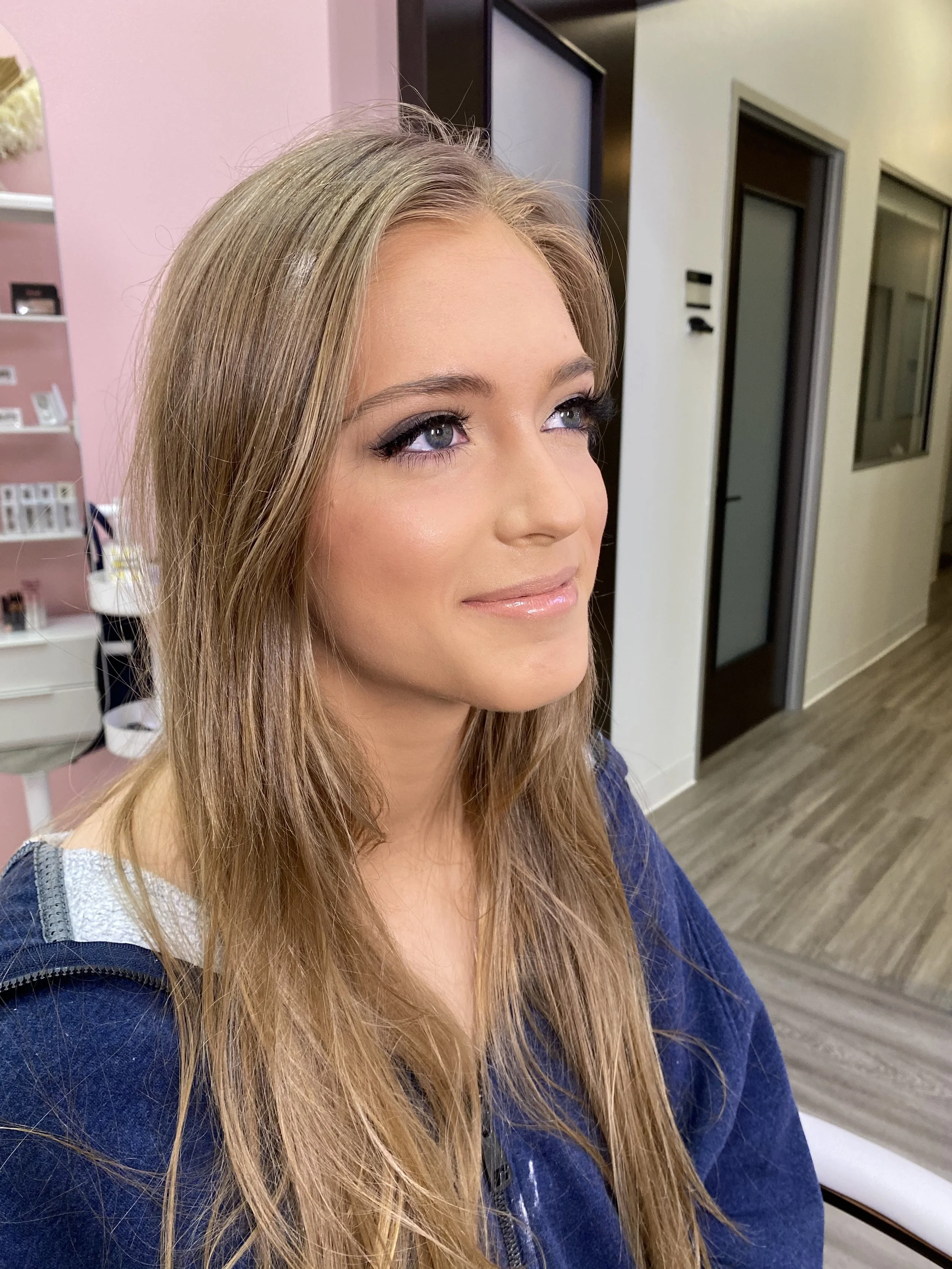 A young woman with long, wavy blonde hair, blue eyes, and light makeup, smiling softly in an indoor setting.