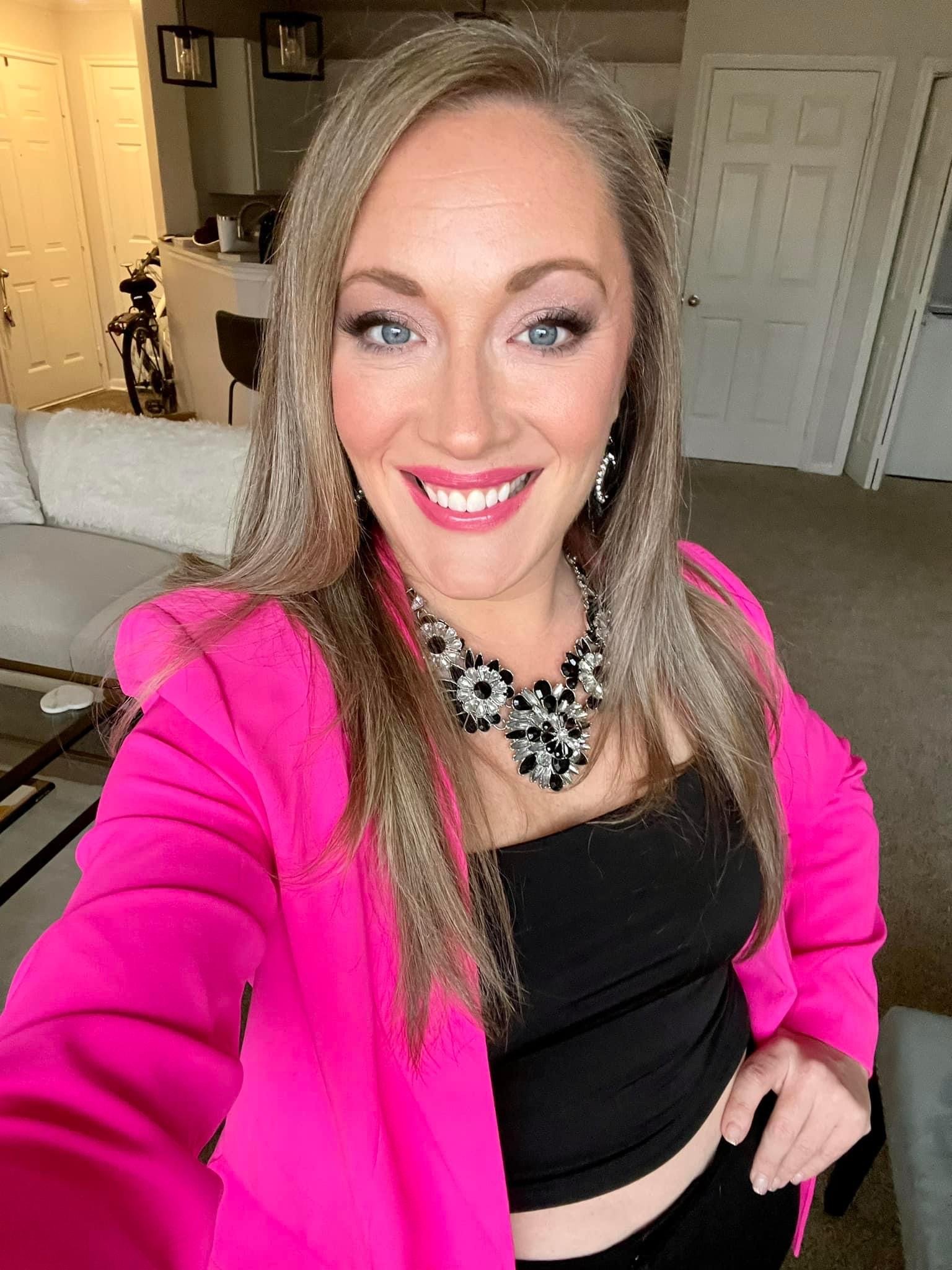 A woman with long blonde hair and blue eyes taking a selfie inside a home. She is wearing a pink blazer, a black top, a large black and silver statement necklace, and matching earrings. The background shows a living room with a beige sofa, a coffee t
