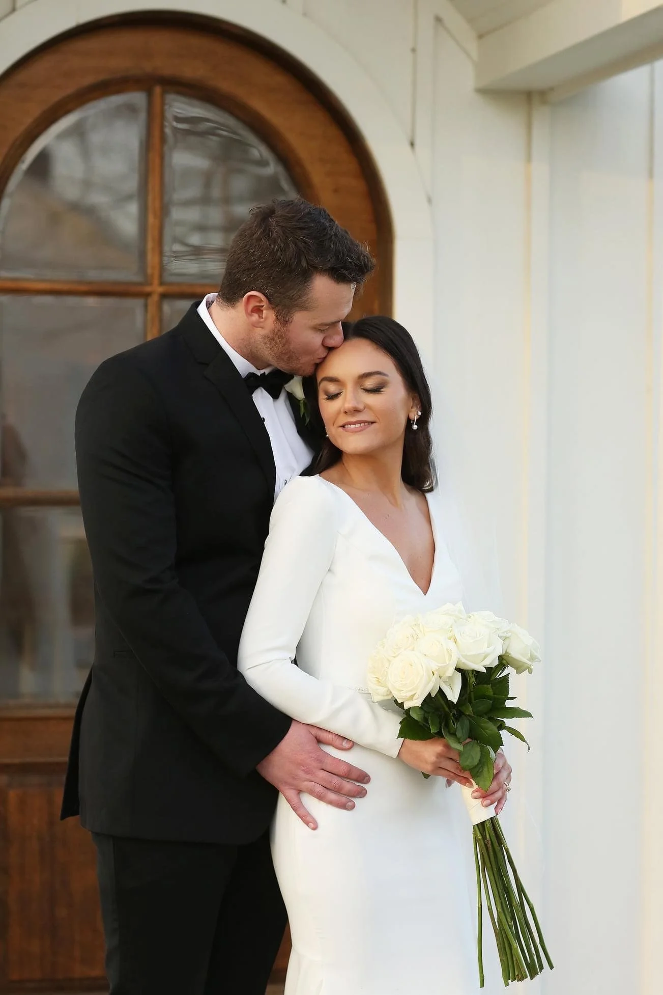 A bride in a white wedding dress holding a bouquet of white roses, with a groom in a black tuxedo kissing her on the forehead, standing in front of a window with a wooden frame.