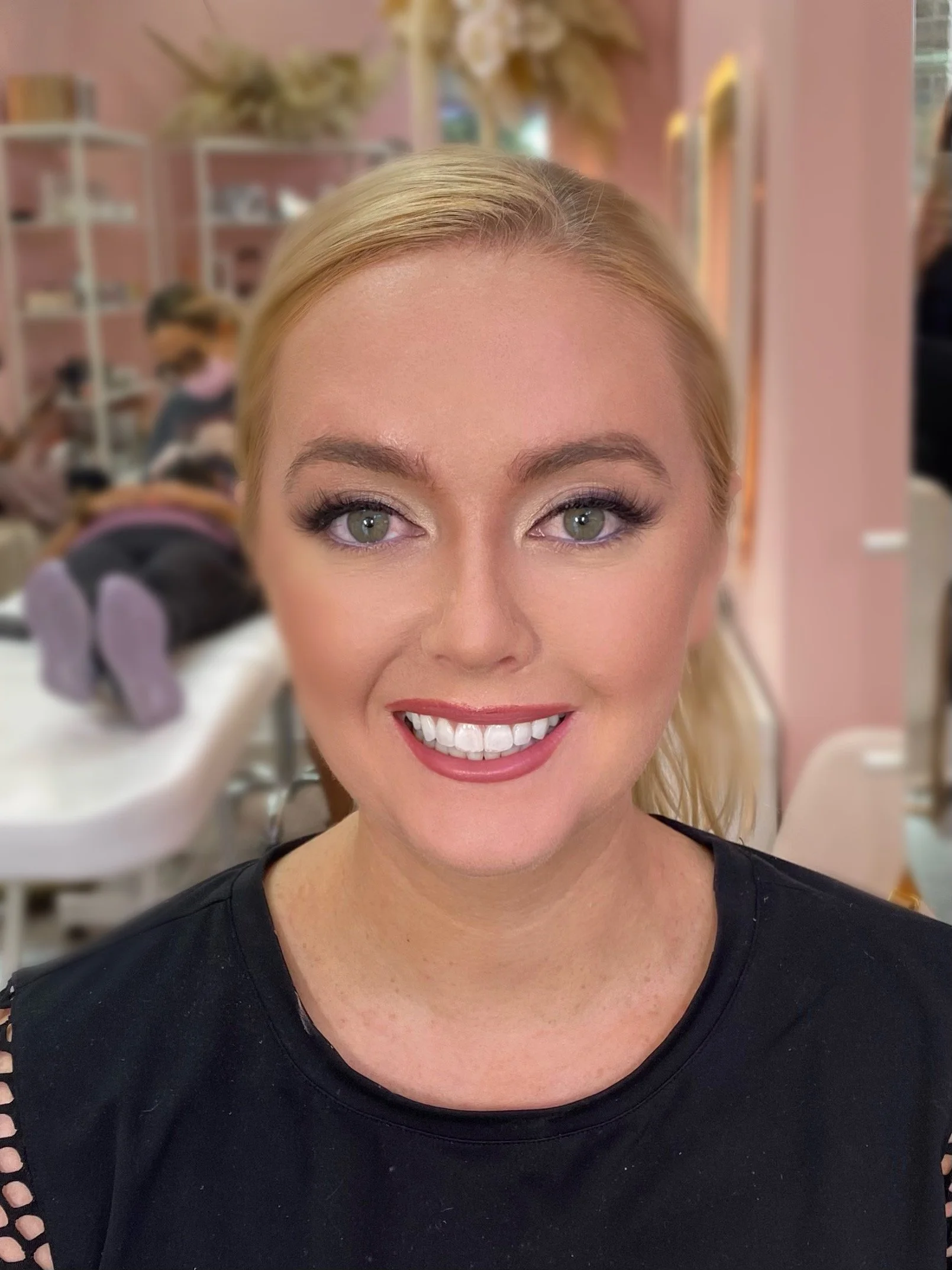 A woman with blonde hair and makeup smiling in a salon or beauty studio, with a person in the background getting a manicure.