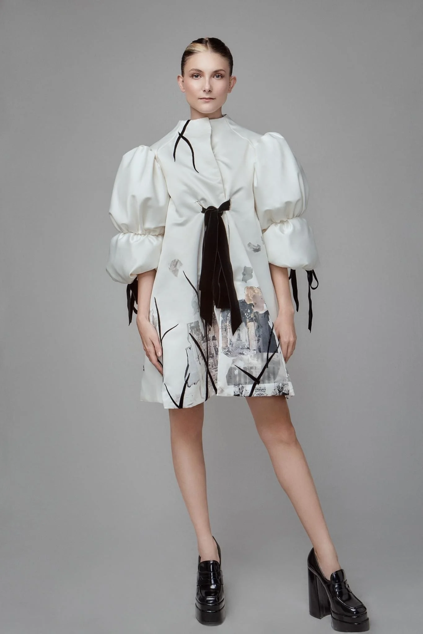 A woman in a fashion-forward dress with puffy, long-sleeved sleeves, black accents, a black ribbon tie at the waist, and an artistic, abstract pattern on the skirt, paired with black platform shoes.