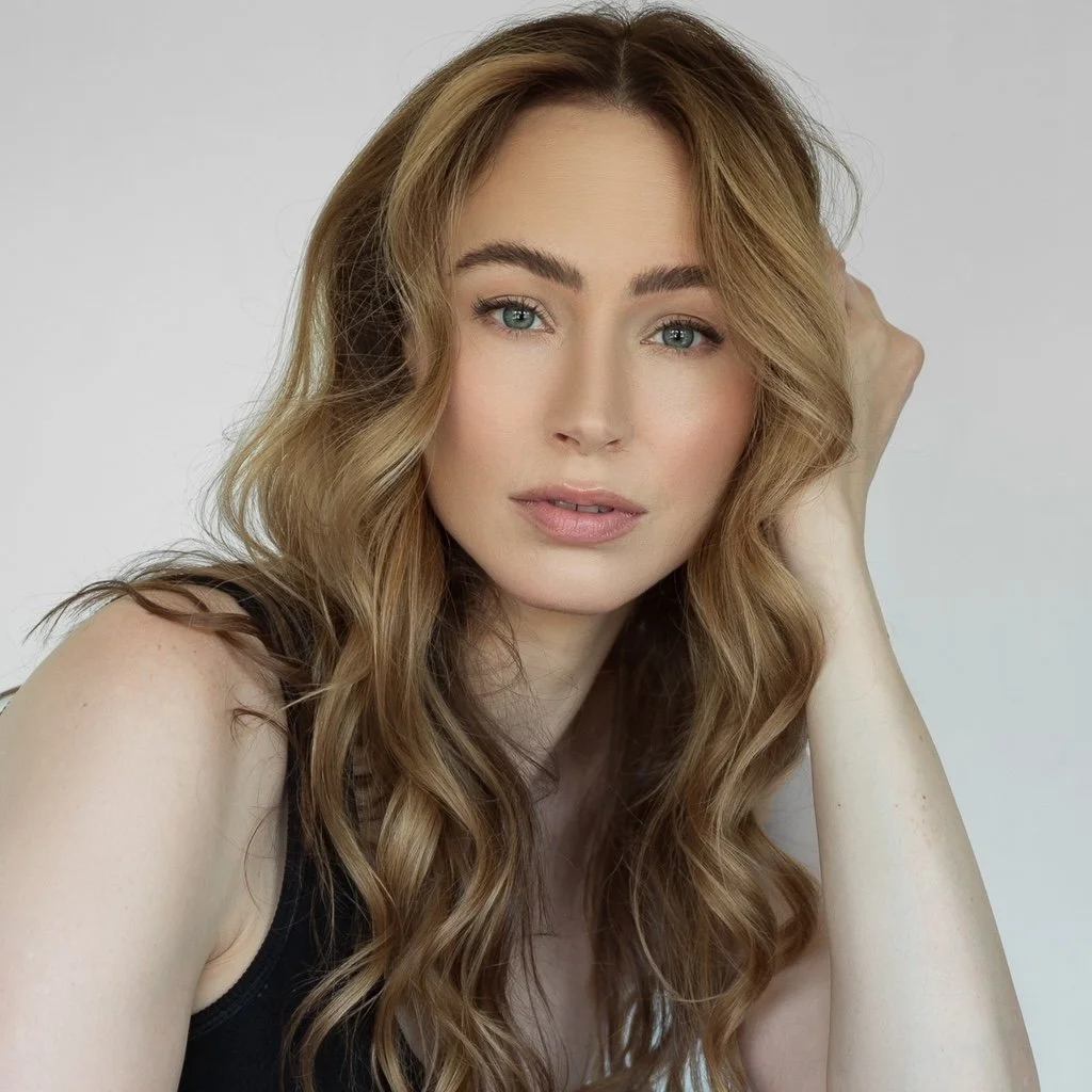 Close-up of a woman with wavy, light brown hair, light makeup, and blue eyes, wearing a black top, posing with her hand resting on her head against a neutral background.