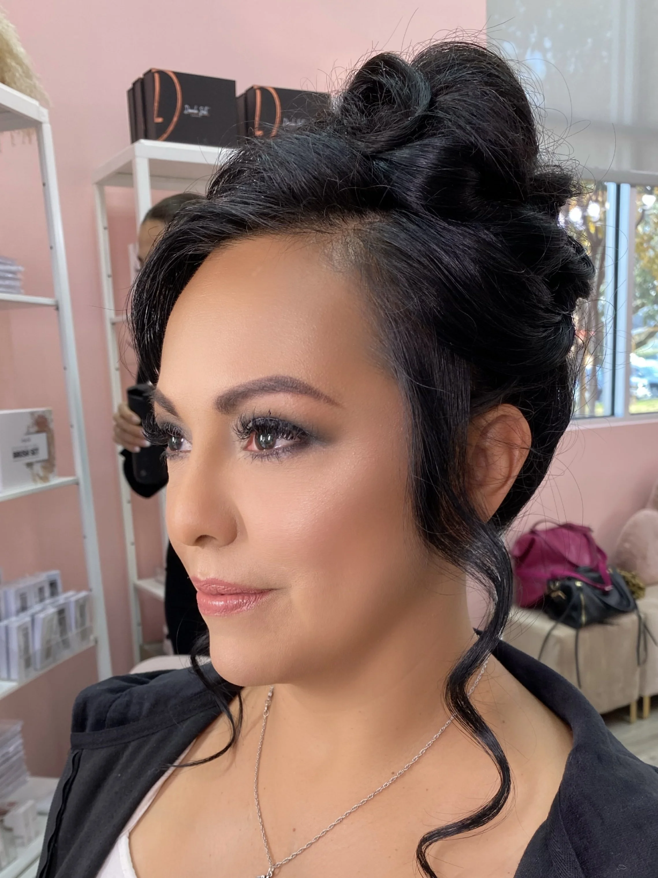 Close-up of a woman with styled black hair, makeup, and a necklace, in a salon or makeup studio setting.