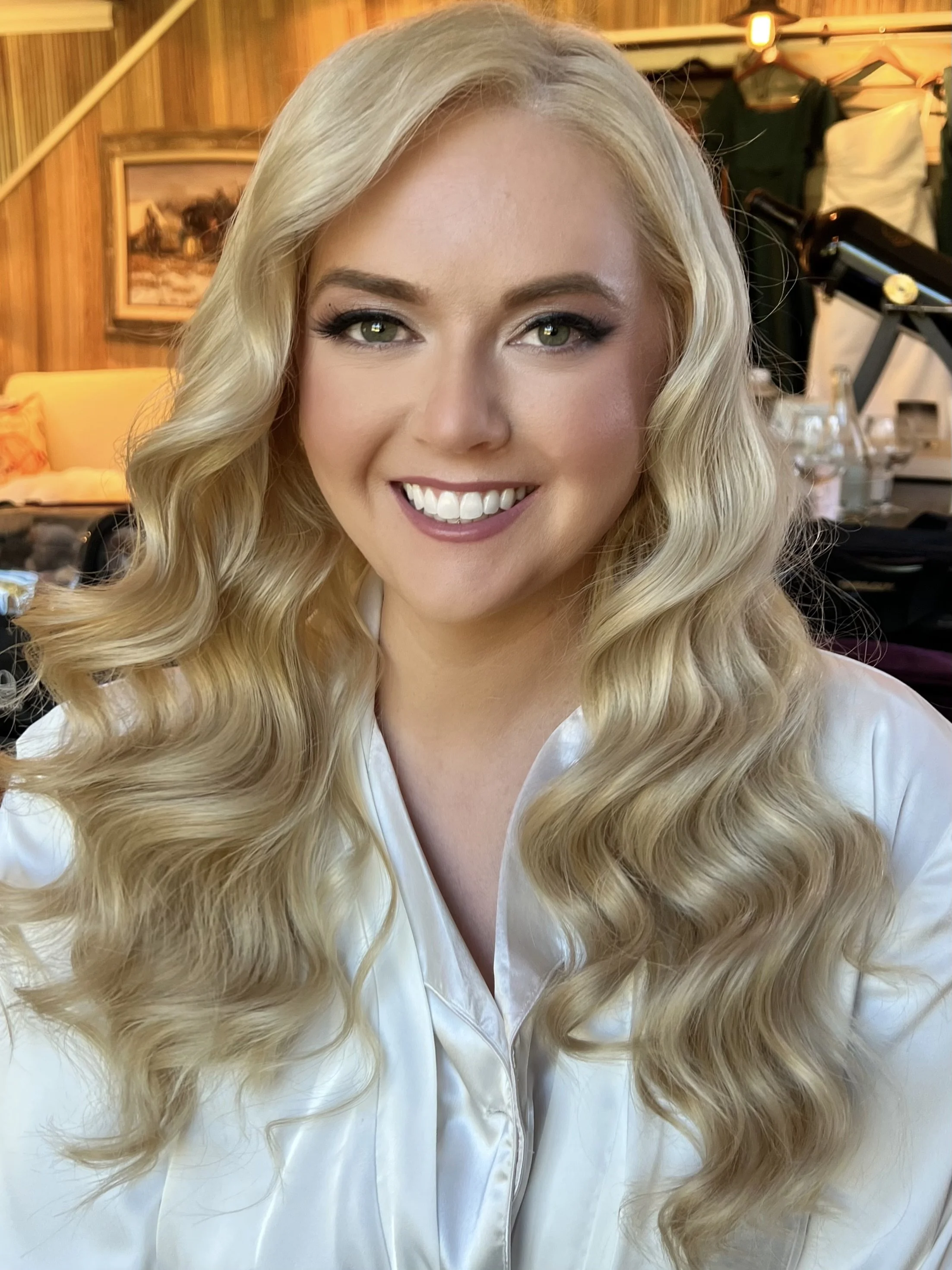 A woman with long, blonde, wavy hair smiling in a setting with wooden walls and framed pictures, wearing a satin white top.
