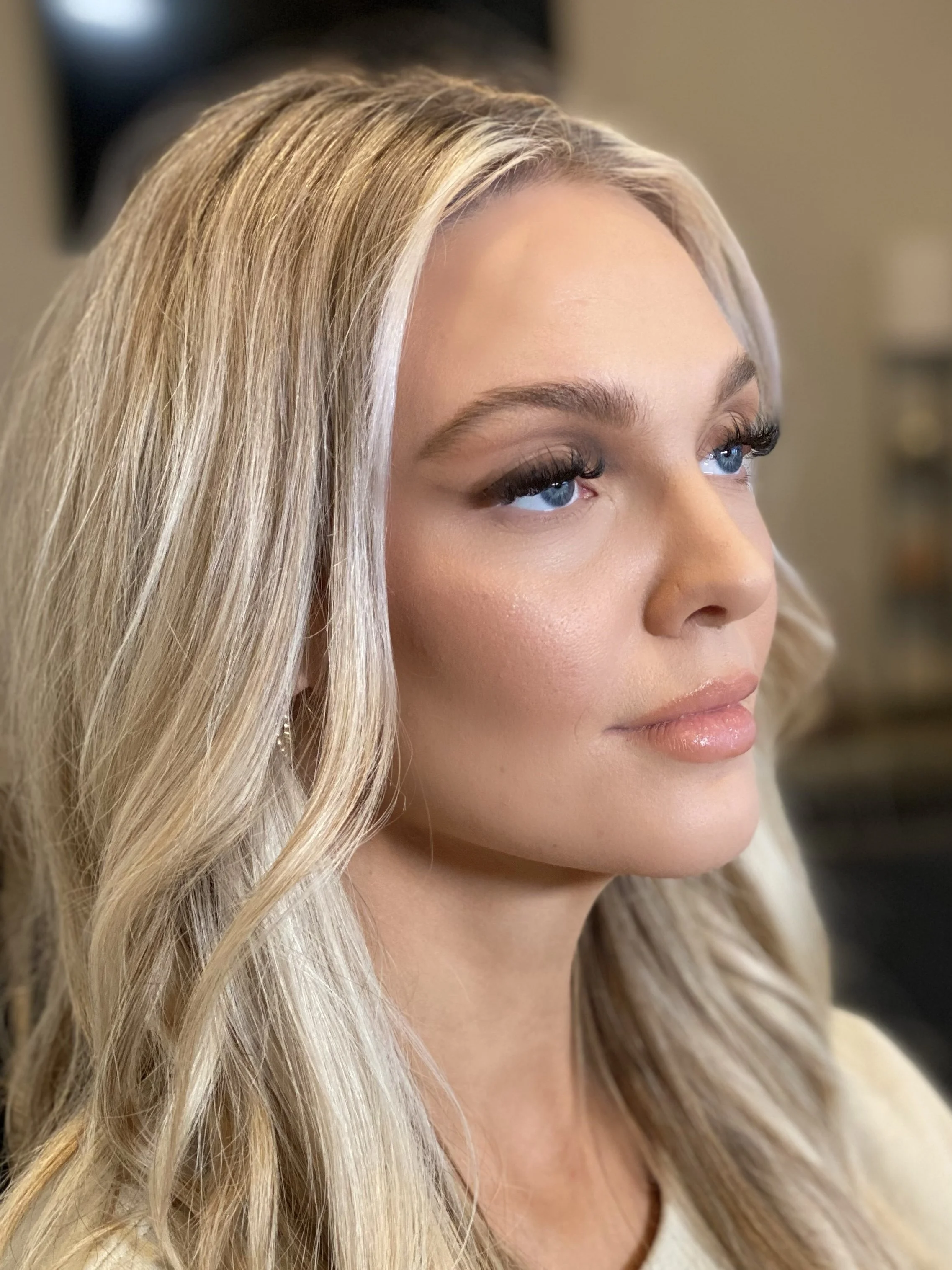 Close-up of a blonde woman with blue eyes, wearing makeup with long eyelashes, glossy lips, and soft waves in her hair, indoors.