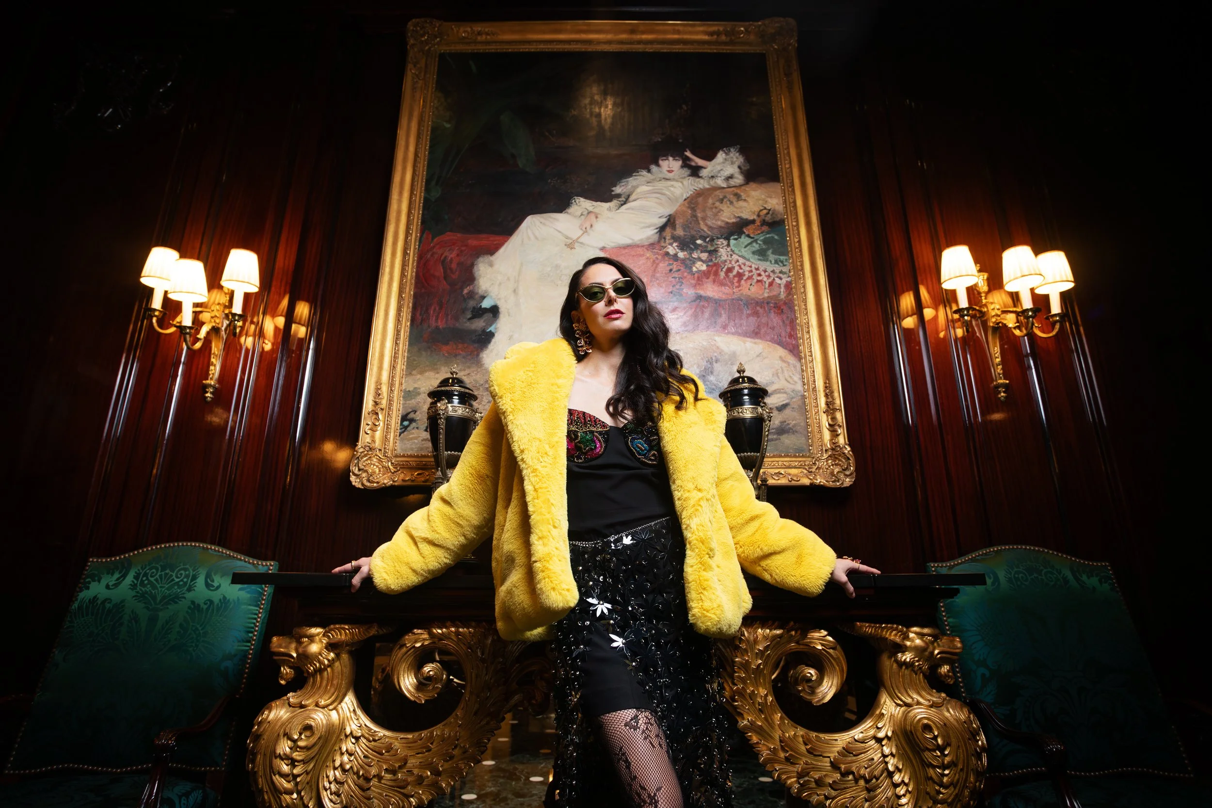 Woman in a yellow fur coat and black dress with floral embroidery, standing in a luxurious room with dark wood paneling, gold accents, green chairs, and a large portrait painting behind her.