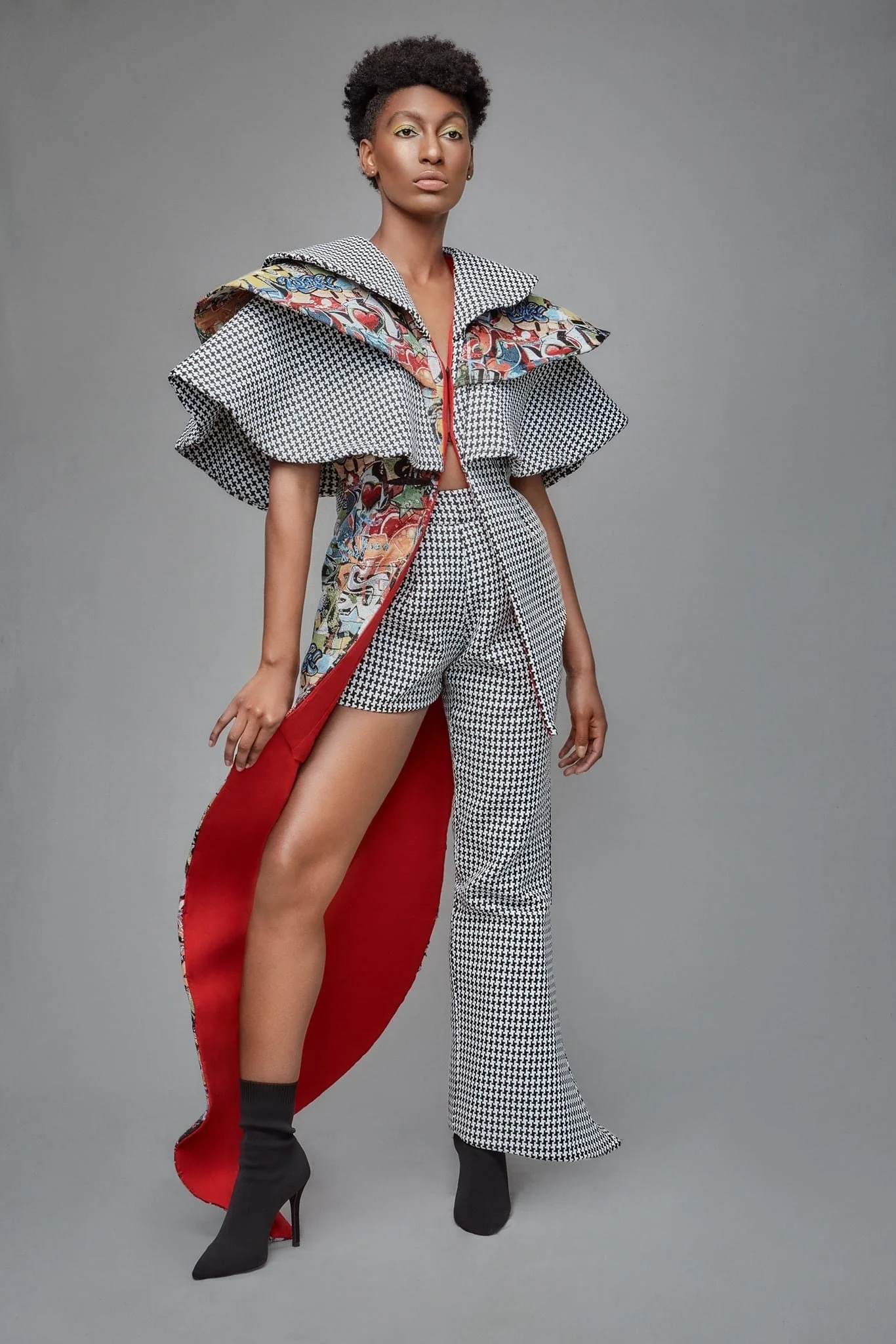 A woman wearing a high-fashion outfit with houndstooth and floral patterns, including wide-leg pants, a structured top with ruffled and floral details, and black high-heeled boots, stands against a grey background.