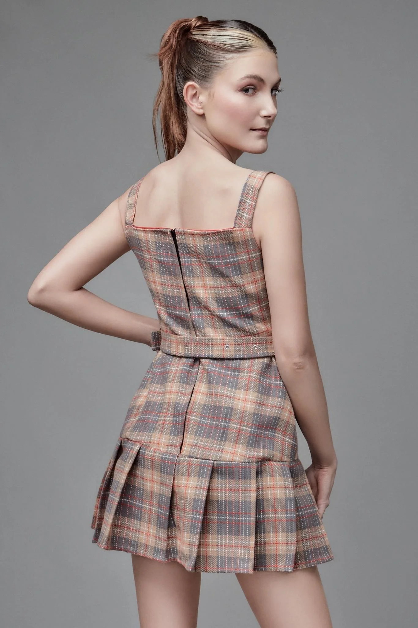 A young woman with styled hair wearing a plaid dress with flared skirt and sleeveless straps, standing against a plain gray background and looking over her shoulder.