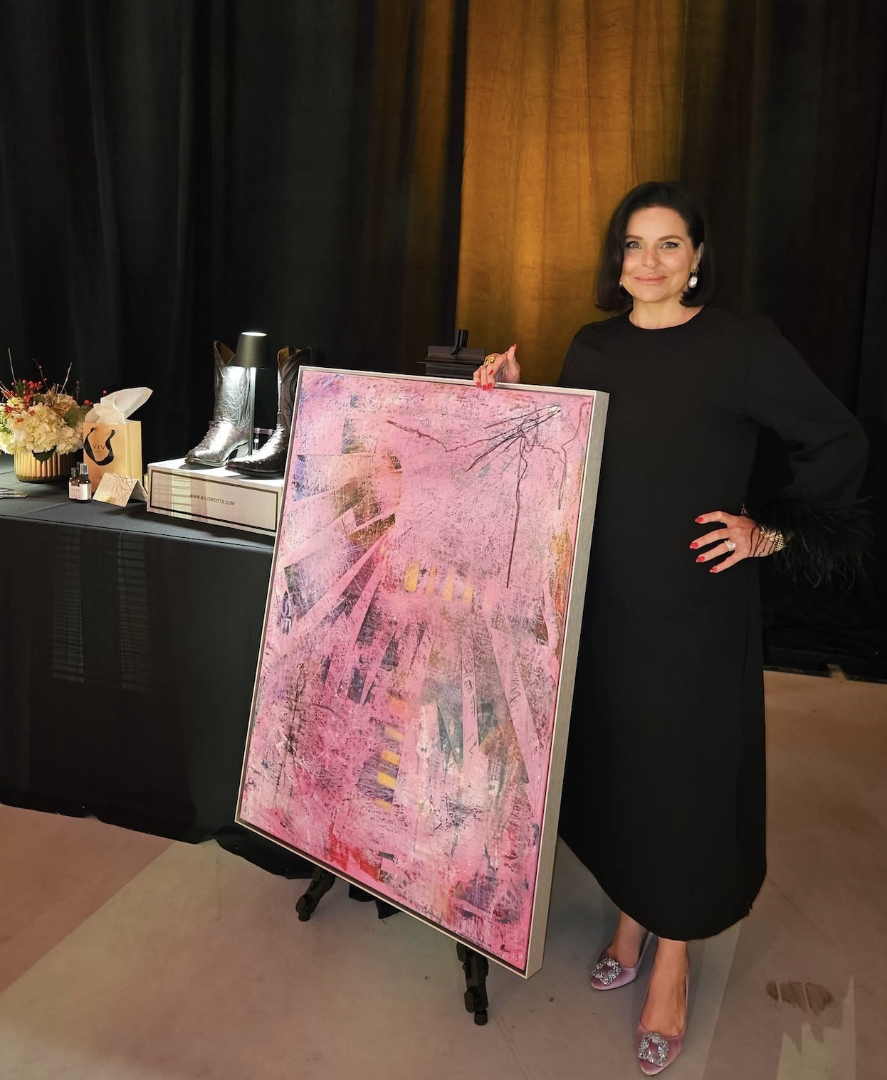 A woman in a black dress standing next to a pink abstract painting on an easel, with a table of shoes and decorative items in the background.