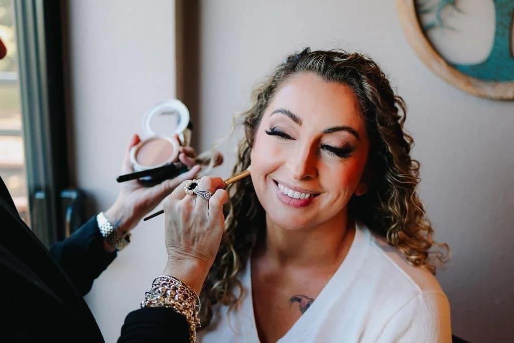 A woman with curly hair getting her makeup done by a makeup artist holding a brush and compact mirror. The woman is smiling with her eyes closed.