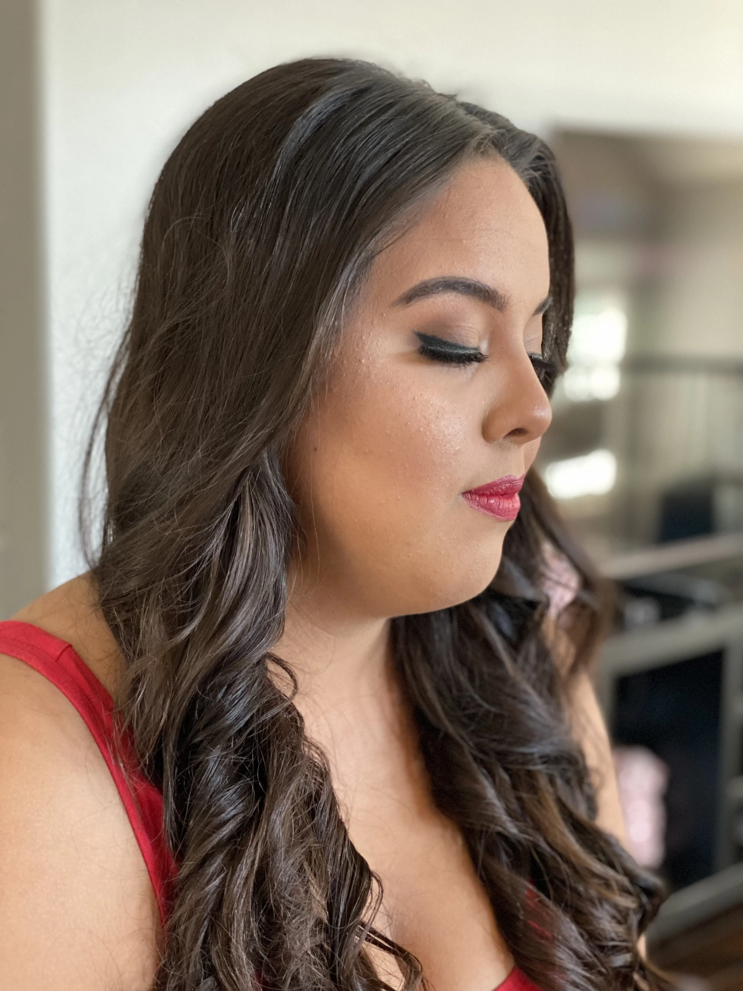 A woman with long curly brown hair and makeup wearing a red top, looking down.