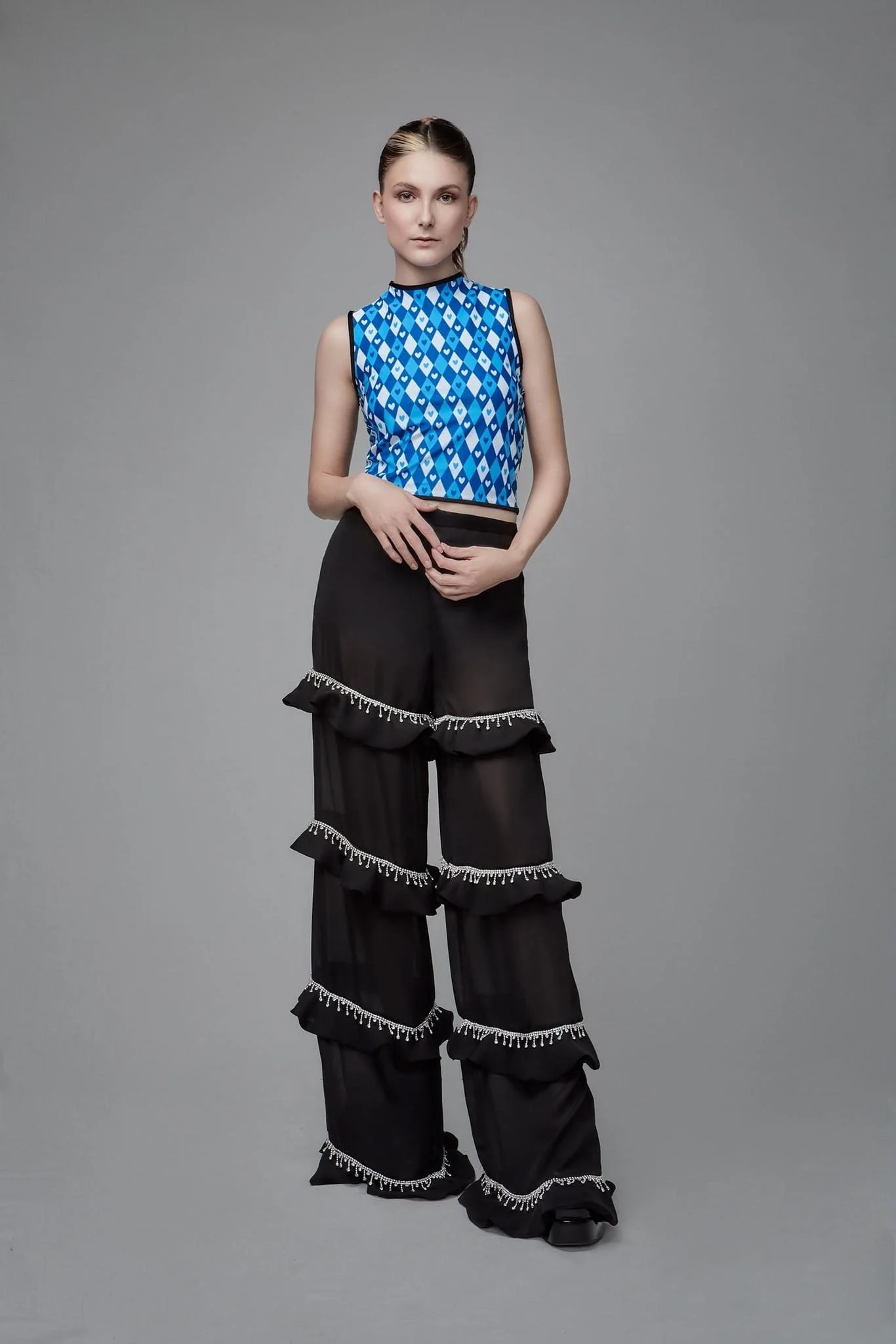 A young woman with brown hair styled back, wearing a sleeveless blue patterned top and black wide-legged pants with ruffles and white decorative trim, standing against a plain gray background.