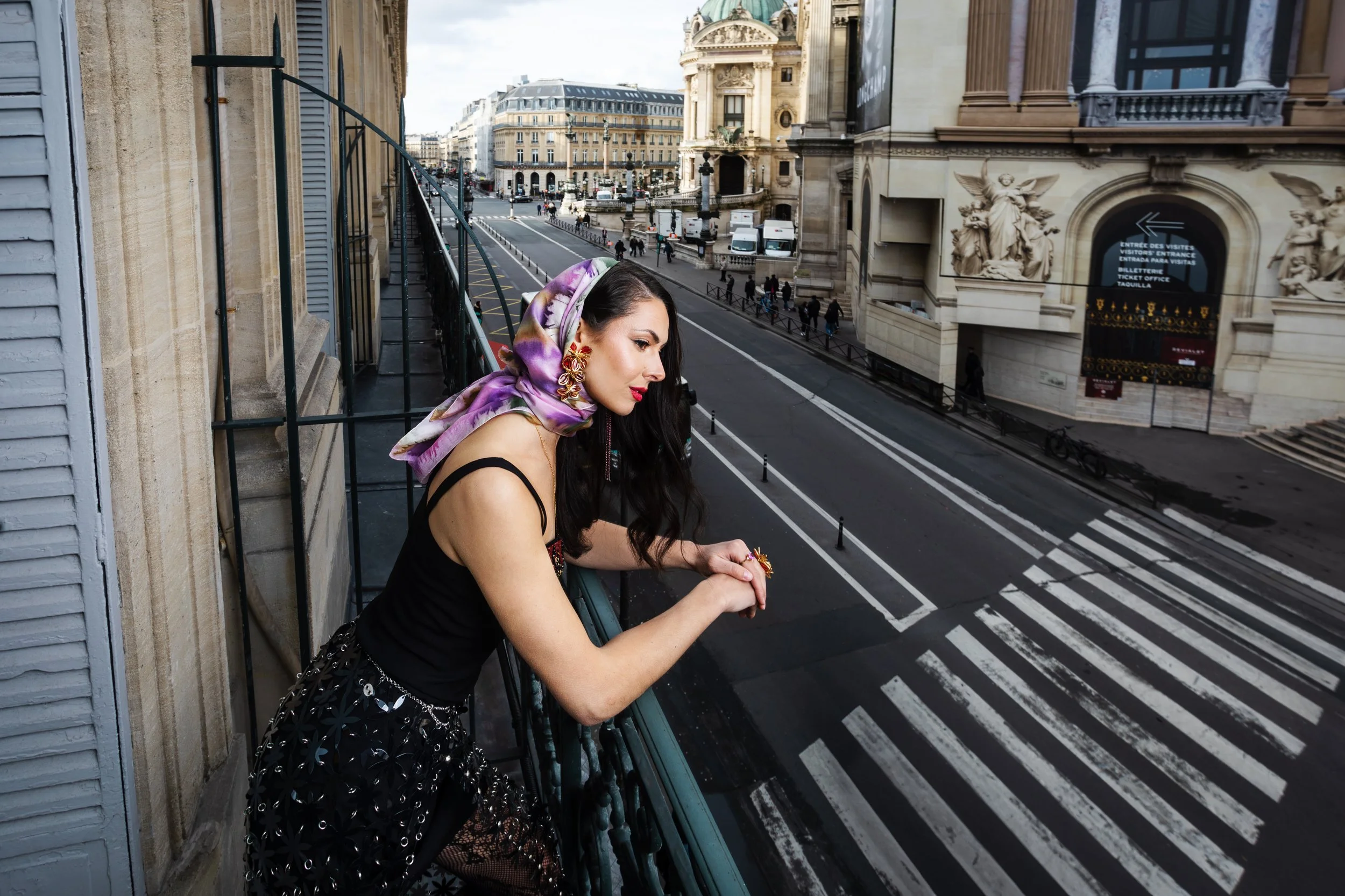 A woman with long dark hair, wearing a black dress and a purple floral headscarf, stands on a balcony overlooking a city street. She has red lipstick, large earrings, and bracelets, leaning on the railing and gazing down.