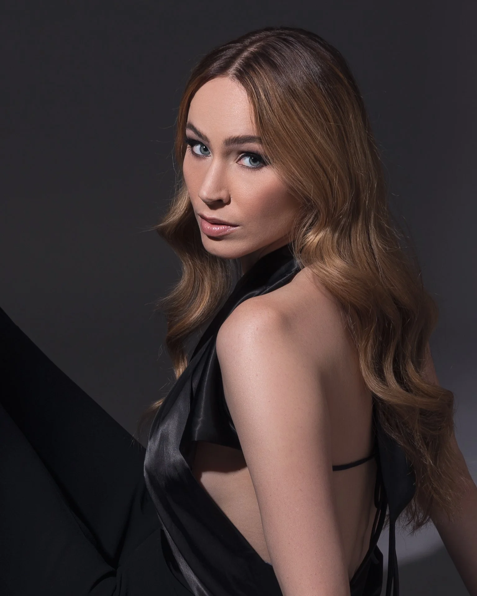 A woman with long wavy hair, looking at the camera with a neutral expression, wearing a black satin top with an open shoulder design against a dark background.