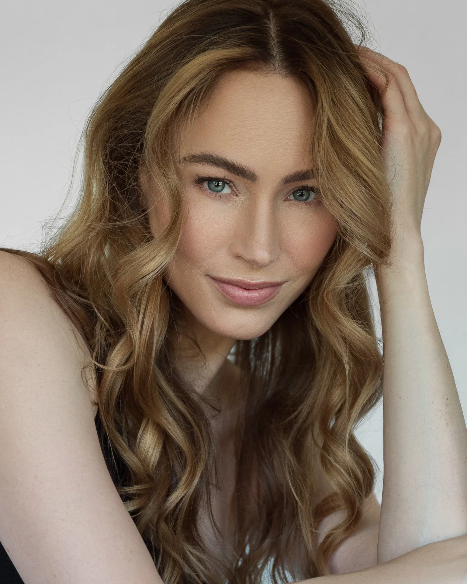 A young woman with long, wavy red hair and blue eyes, smiling softly, with her hand resting on her head.