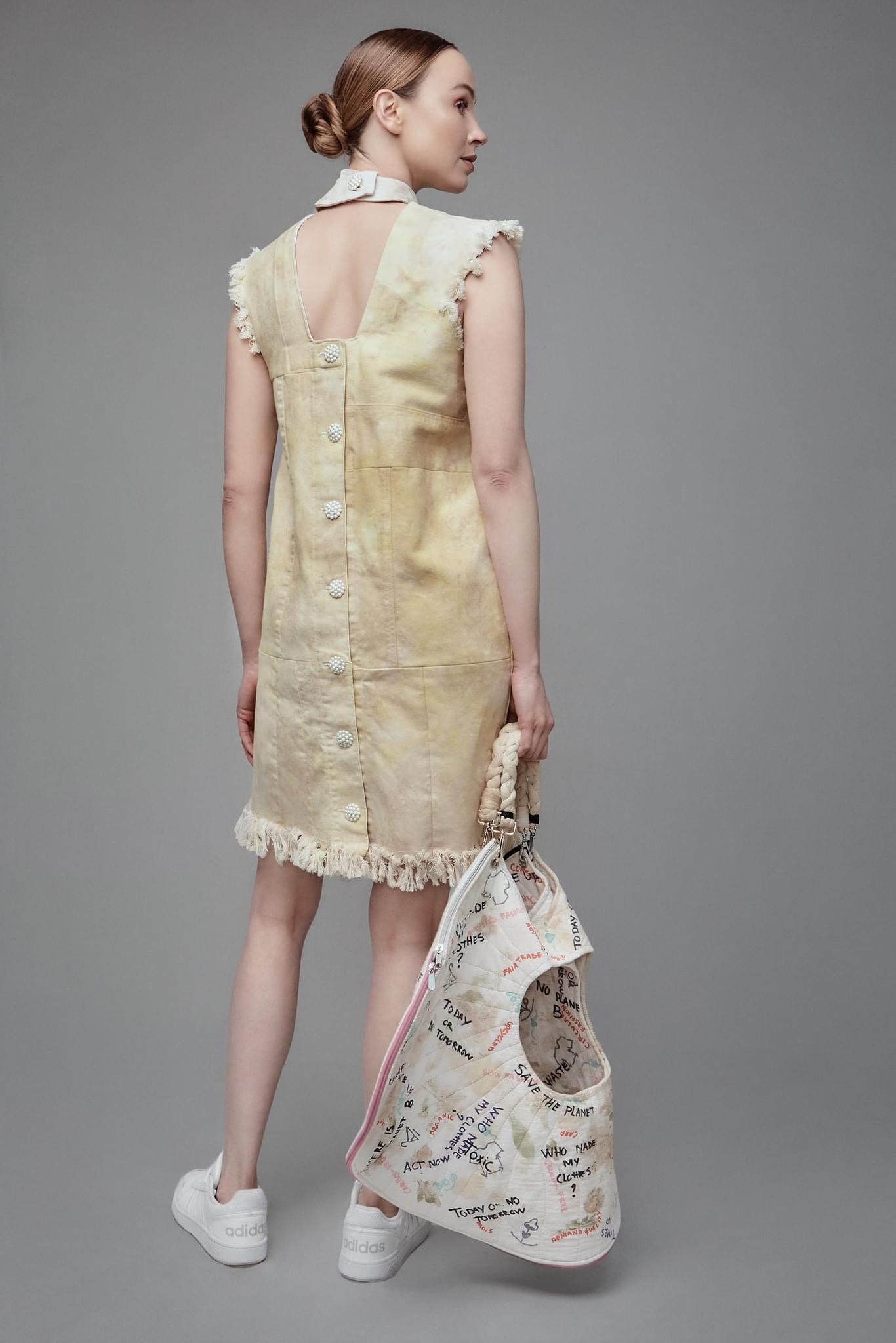 Fashion model wearing a sleeveless beige dress with frayed edges, holding a large tote with colorful writing and graphics, white sneakers, and a white scarf tied around her neck, standing against a plain grey background.