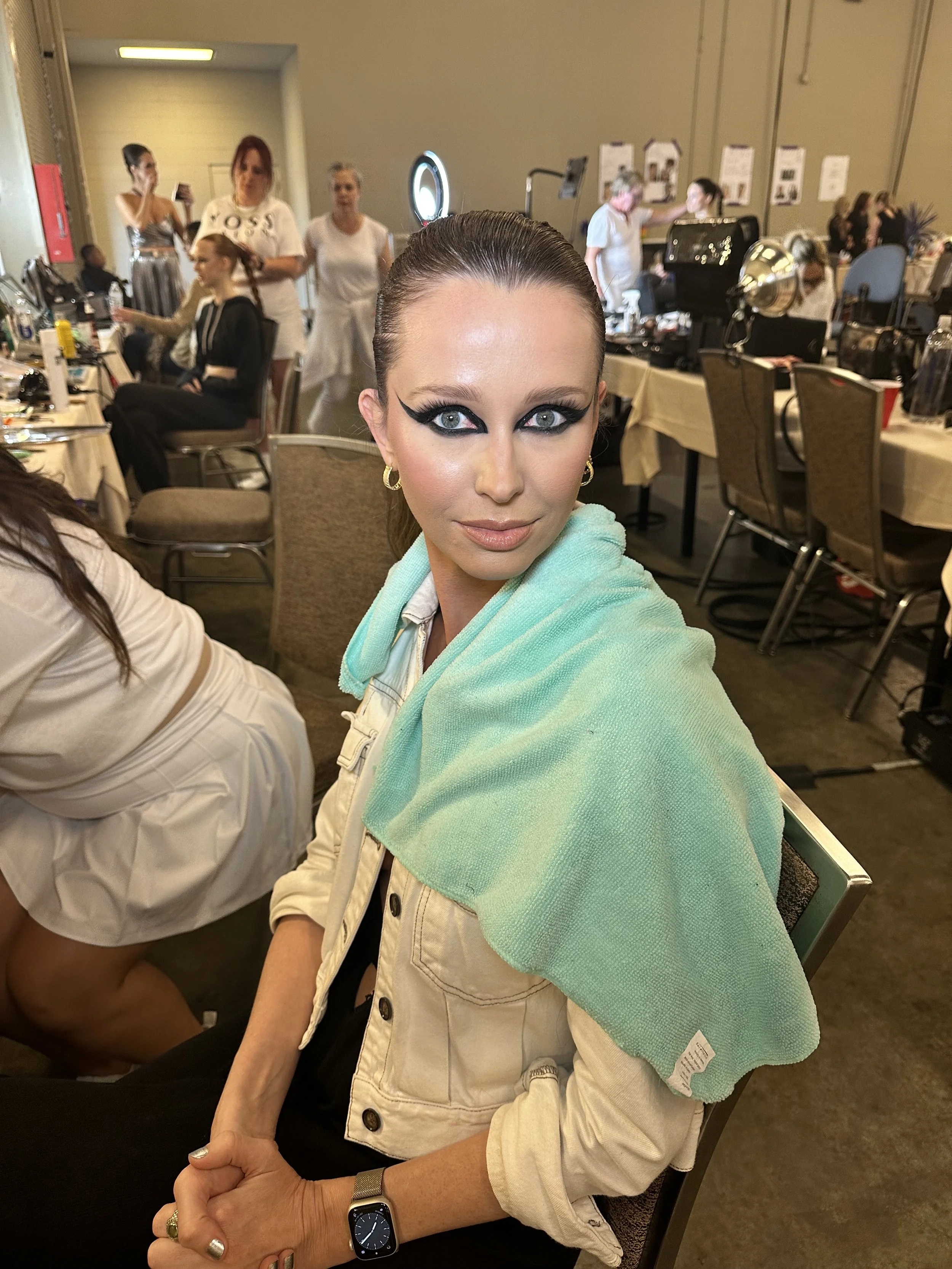 A woman with dramatic black eye makeup and sleek hair is sitting in a backstage area, with makeup and hairstyling tools visible around her and other people preparing in the background.