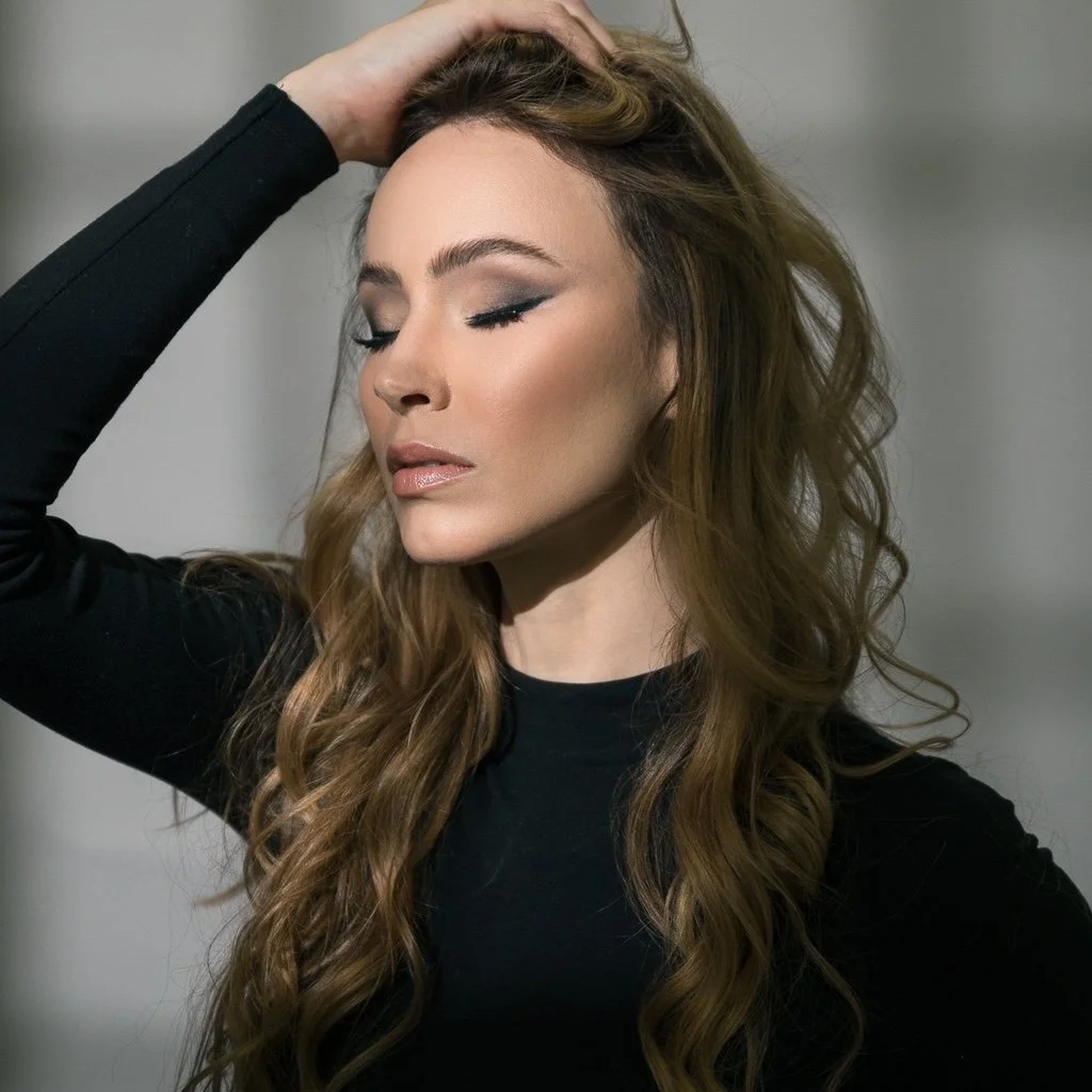 A woman with long wavy brown hair and makeup, wearing a black top, with her eyes closed and one hand resting on her head.