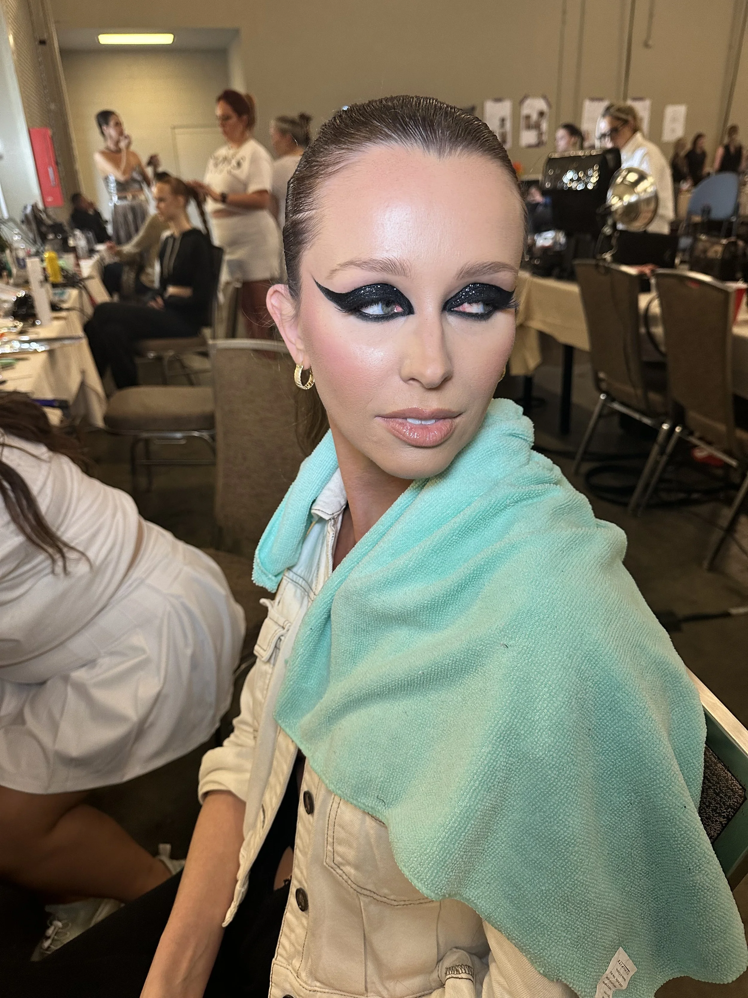 Woman with elaborate black eye makeup and shiny black eyeshadow, wearing gold hoop earrings, with a green towel over her shoulder, seated in a backstage area with people and makeup stations in the background.
