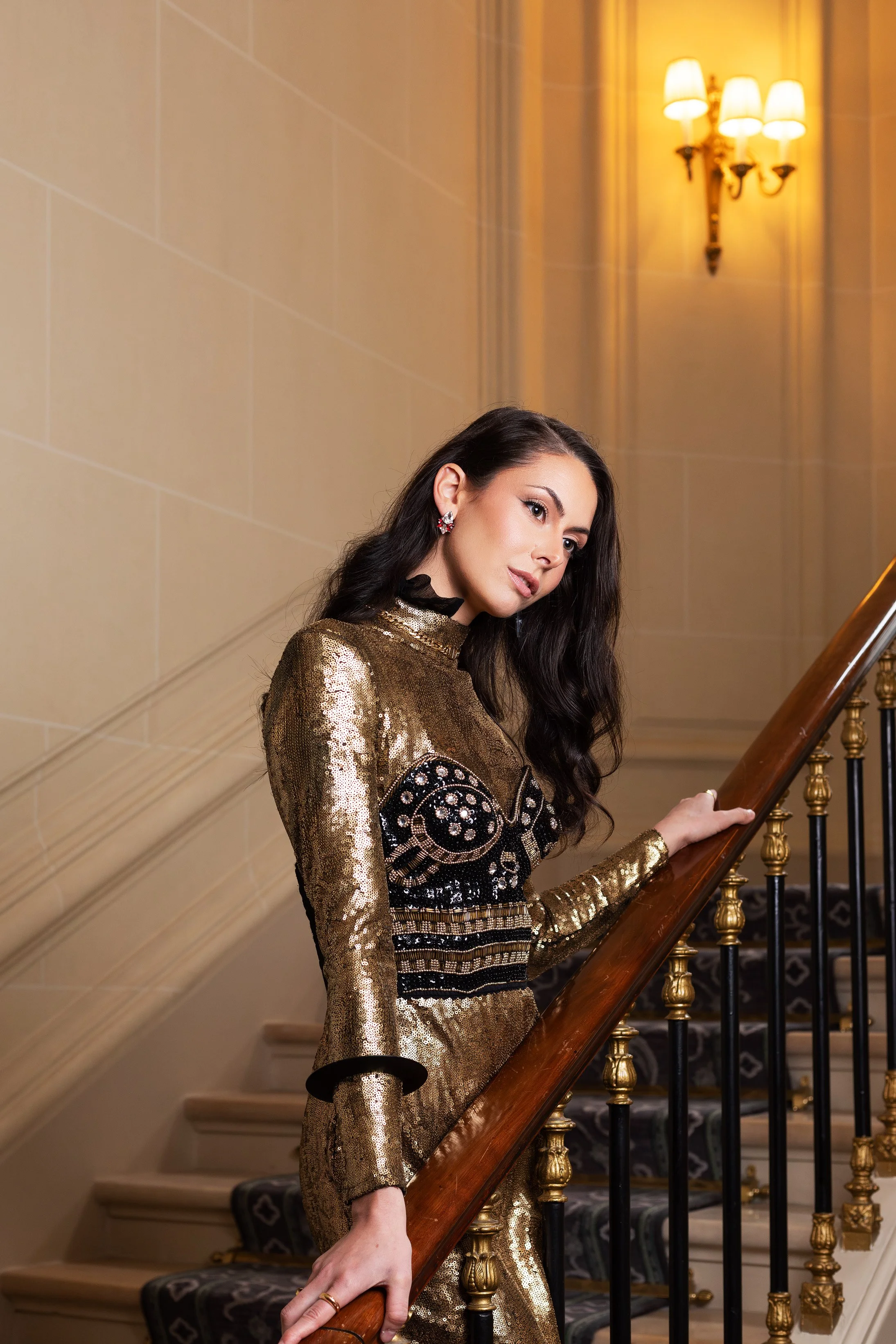 A woman in a shiny gold and black dress is standing on a staircase, holding the wooden handrail, and looking to her right. The setting has warm lighting, with a wall-mounted sconce visible in the background.