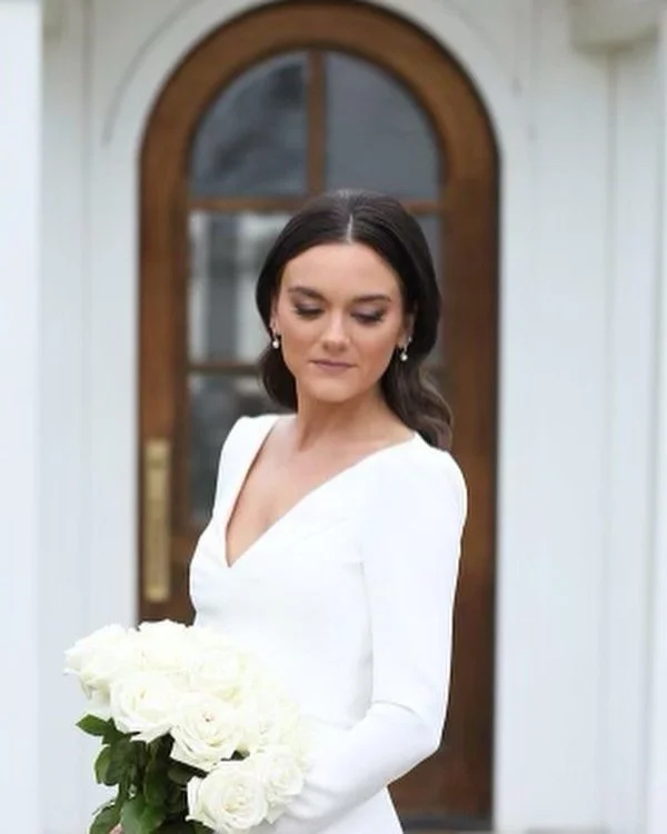 Woman in white dress holding a bouquet of white roses standing in front of a wooden door with an arched window, outdoors.