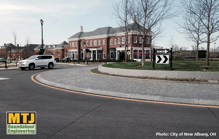 A roundabout with a brick building in the background, leafless trees, and a white SUV navigating the intersection in New Albany, Ohio.