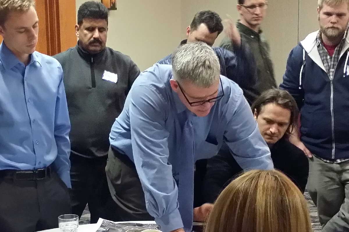 Group of people gathered around a table, with a man in a blue shirt leaning over papers, engaged in discussion.