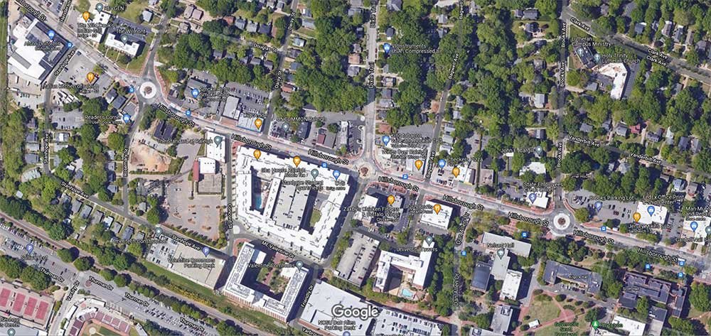 Aerial view of a city neighborhood with buildings, streets, and green spaces, featuring Hillsborough Street in Raleigh, NC, as well as labeled places and points of interest.