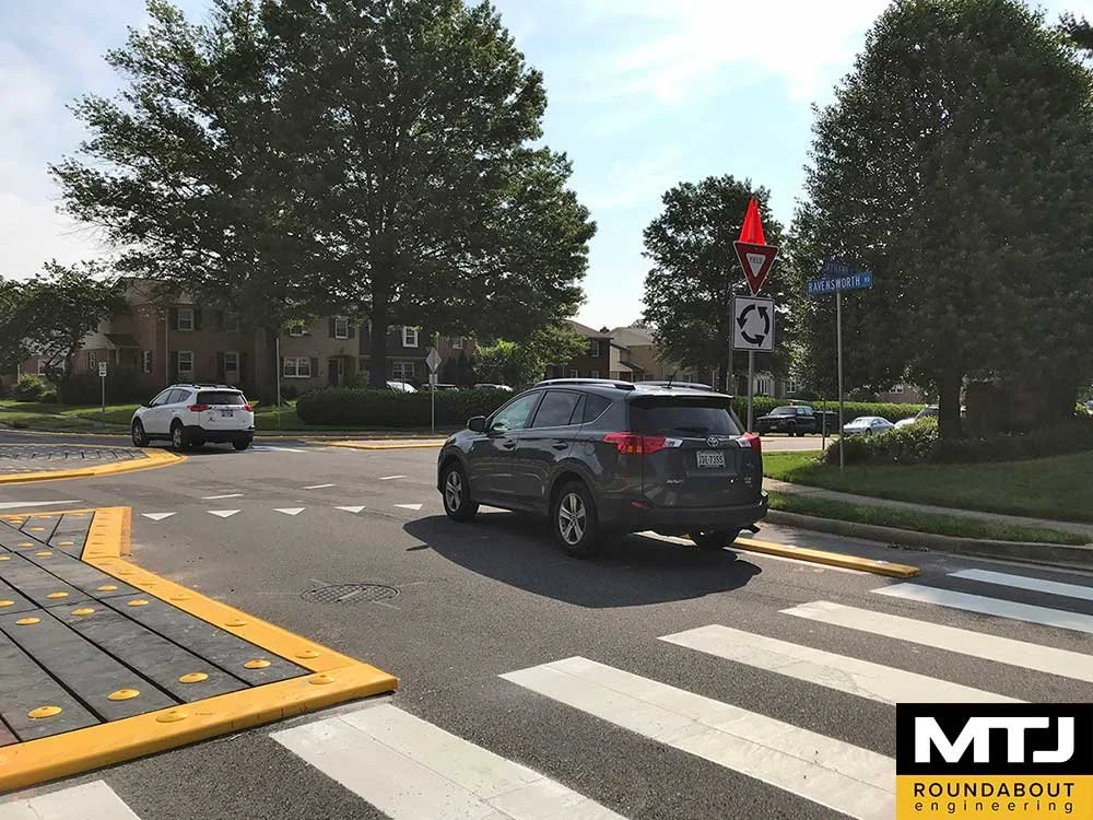 Two cars at a roundabout with clear road markings and trees in the background, "MTJ Roundabout Engineering" logo in the corner.
