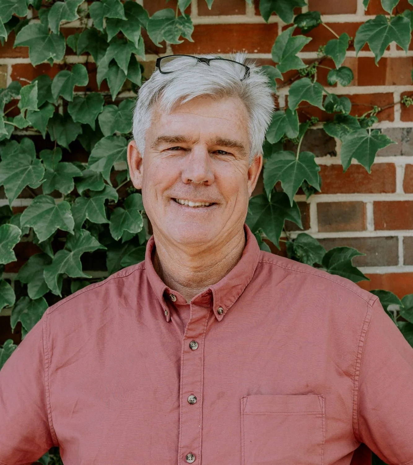 A smiling middle-aged man with gray hair and glasses on his head, wearing a salmon-colored button-up shirt, standing outdoors in front of a brick wall covered with green ivy leaves.
