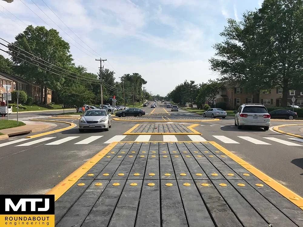 Road with rounded speed bumps, crosswalk, and cars driving, surrounded by trees and houses on a sunny day.