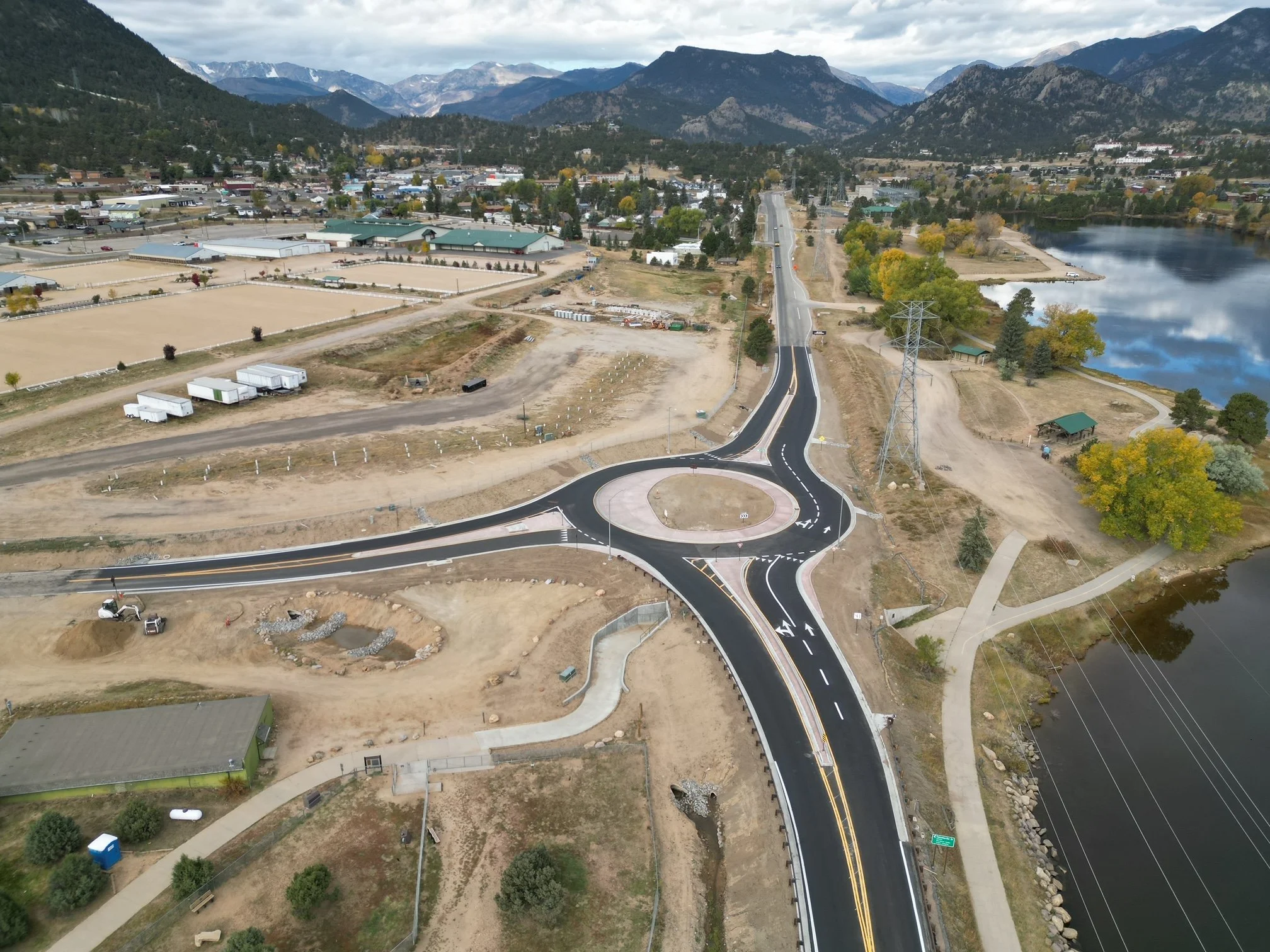 Aerial view of a roundabout with surrounding roads, construction area, and buildings in a town, amidst mountainous landscape.
