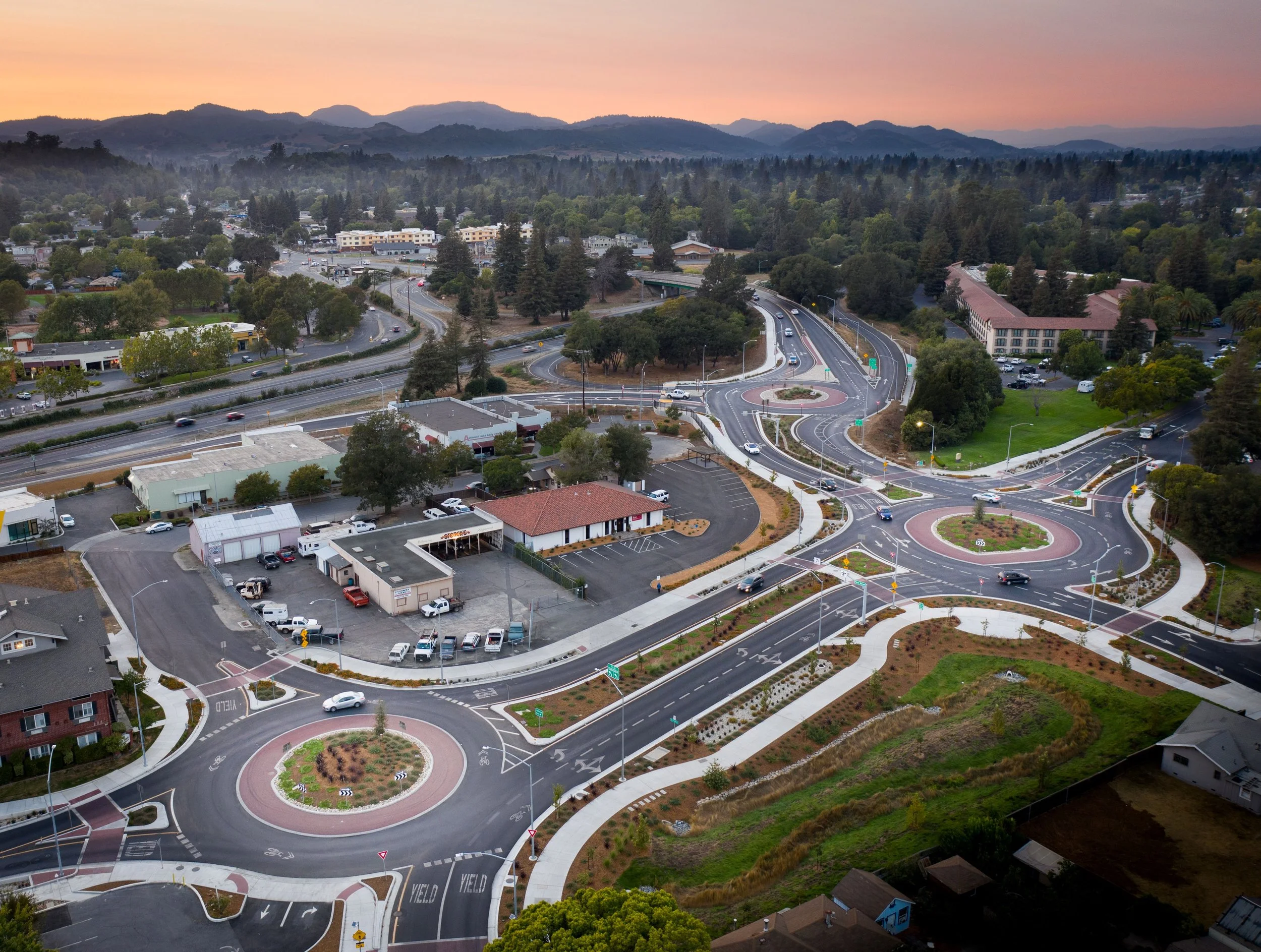 Aerial view of a roundabout with multiple lanes, landscaped islands, and surrounding roads, buildings, trees, and mountains in the distance at sunset.