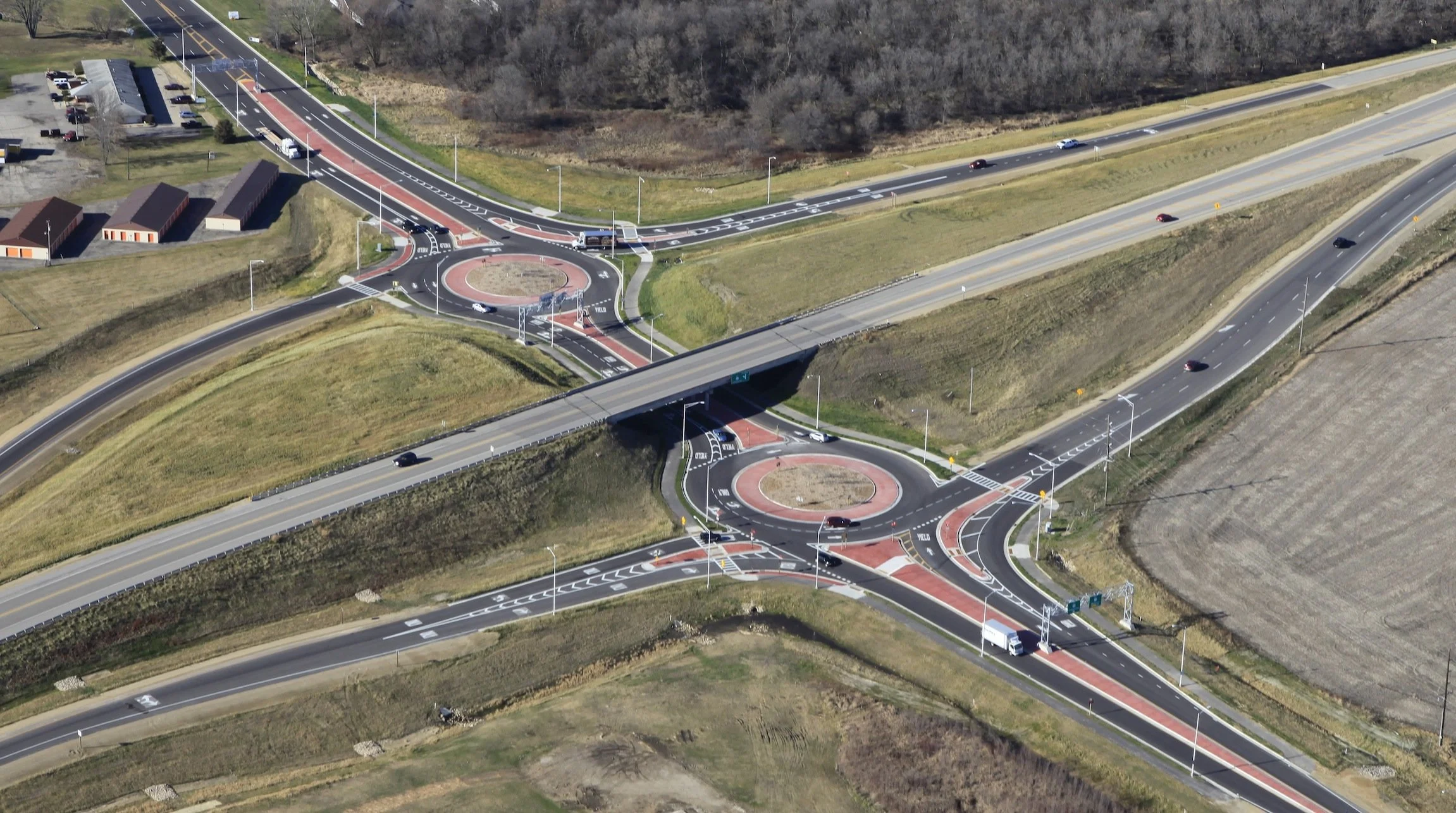 Aerial view of a road interchange with multiple roundabouts, overpasses, and connecting roads, surrounded by fields and some buildings.
