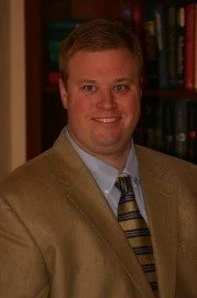A man with short brown hair smiling, wearing a tan blazer, light blue shirt, and striped tie, standing in front of a bookshelf filled with books.