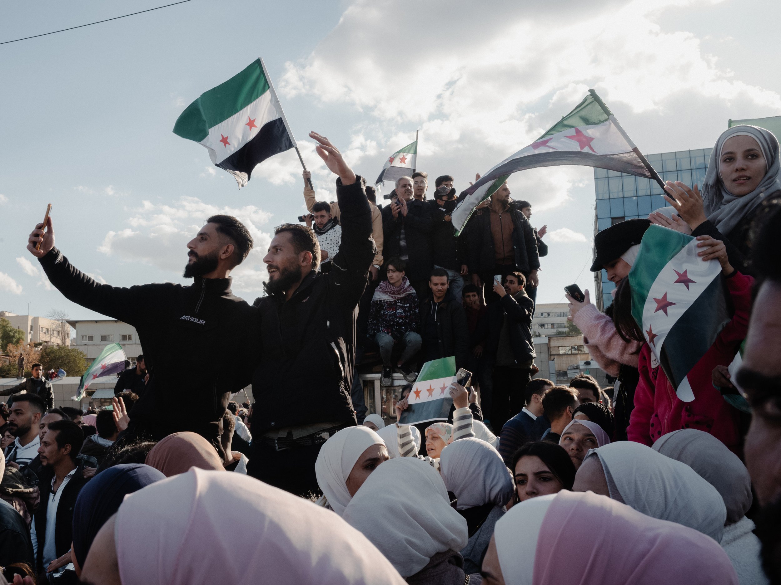 Crowd of people gathered outdoors with some holding flags featuring green, white, black, and red colors, under a partly cloudy sky.
