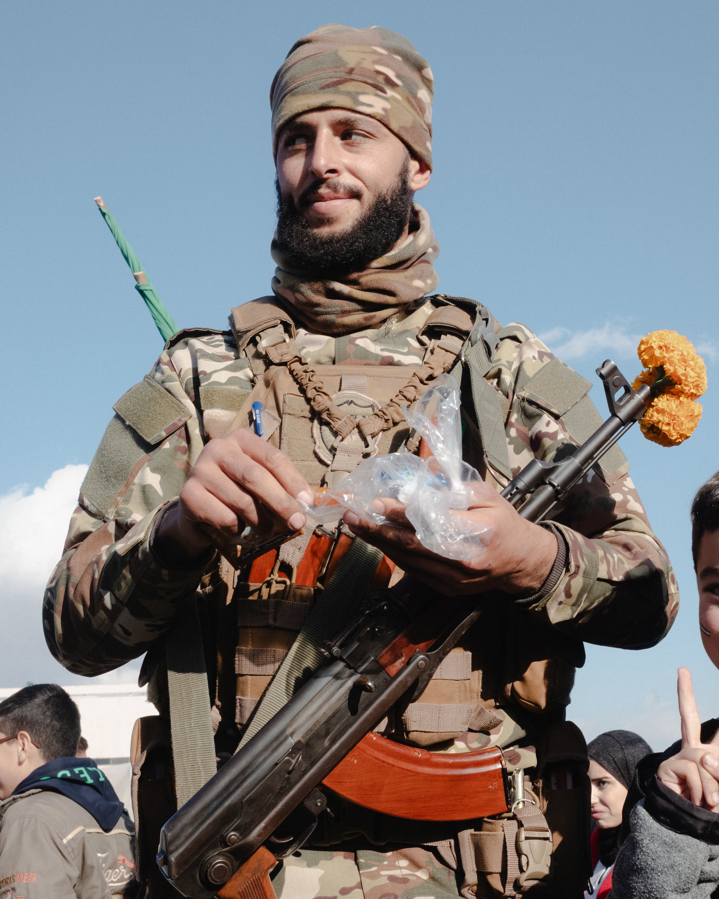 A man in military camouflage gear holding a gun with a yellow marigold flower attached to it, standing outdoors with a clear blue sky and a crowd of people around.