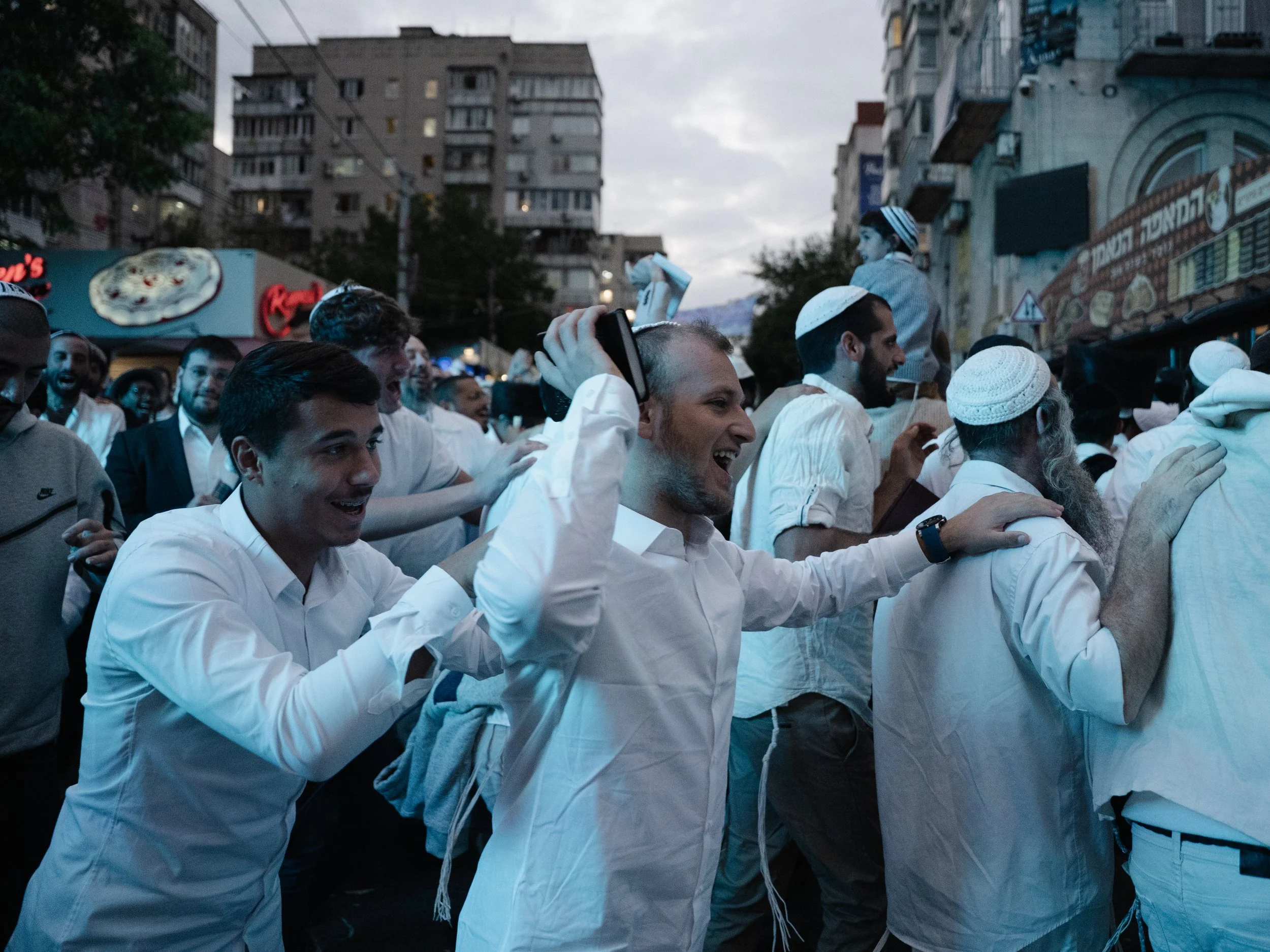 Crowd of people, mostly men, celebrating on a street during dusk, with some wearing white shirts and yarmulkes, in front of buildings with signs including a pizza logo.