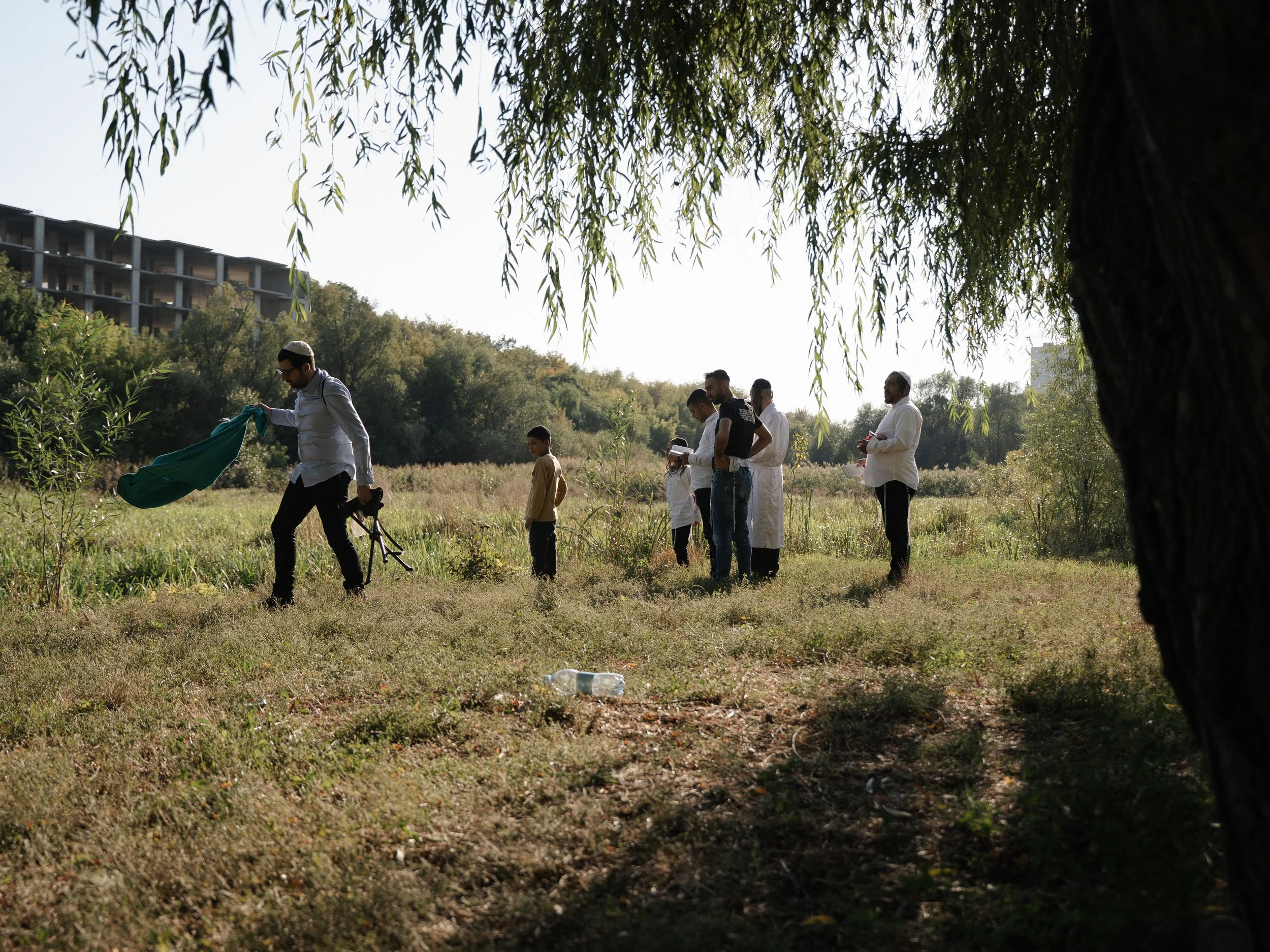 Group of people, including children, gathered outdoors near a large tree in a grassy area, with a building visible in the background.