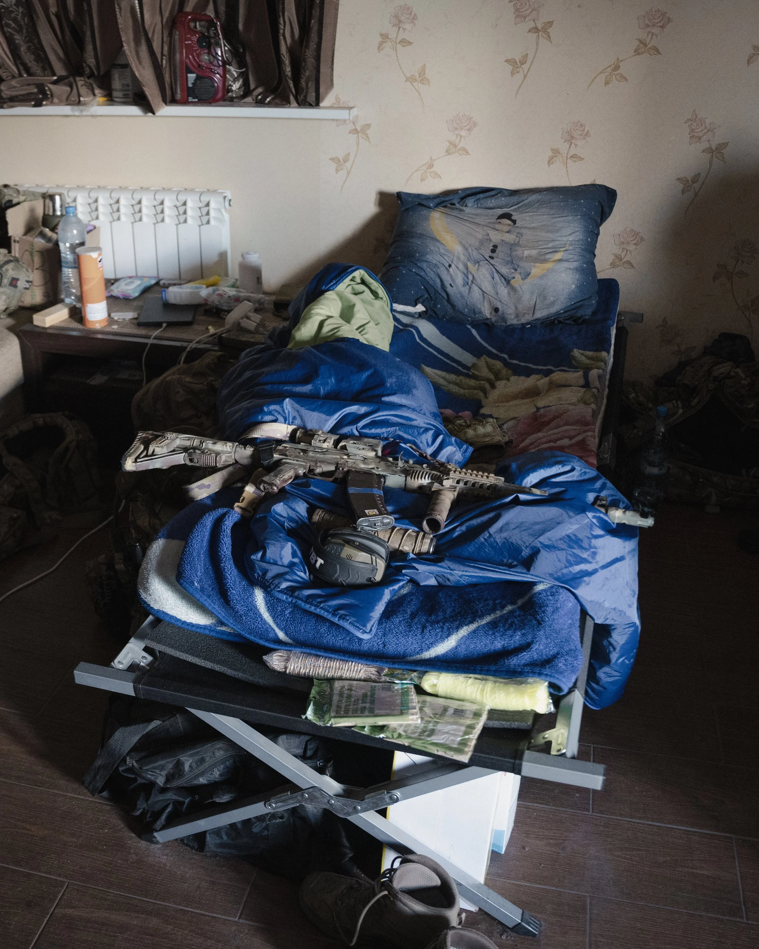 A cluttered hospital bed with disorganized items including clothes, medical equipment, and personal belongings. The bed has a mattress with a nightstand beside it, and there are water bottles, a backpack, a pair of shoes, and various other objects scattered on and around it.