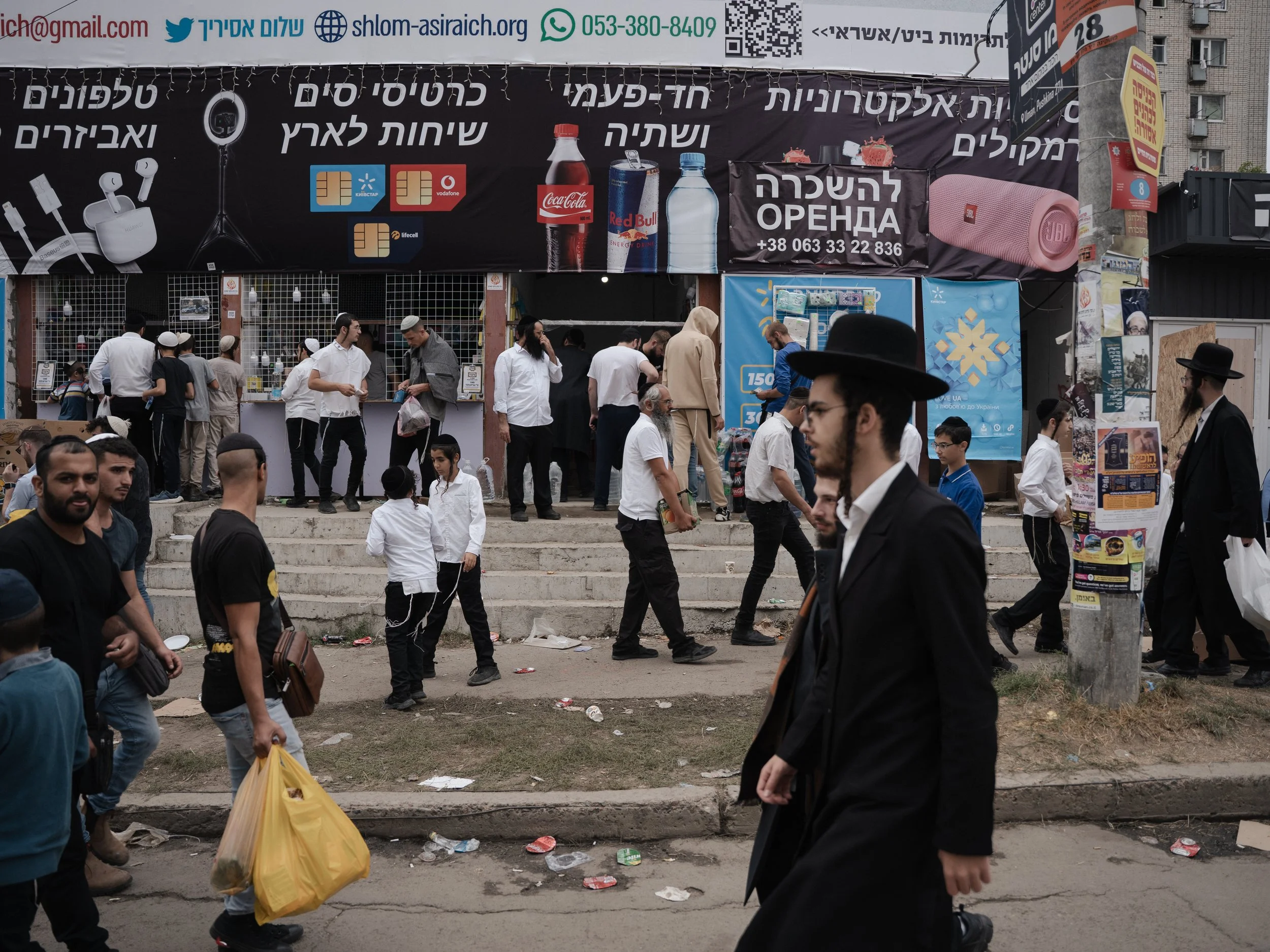 People walking and standing near a building under a large sign in a crowded outdoor area, with trash on the ground.