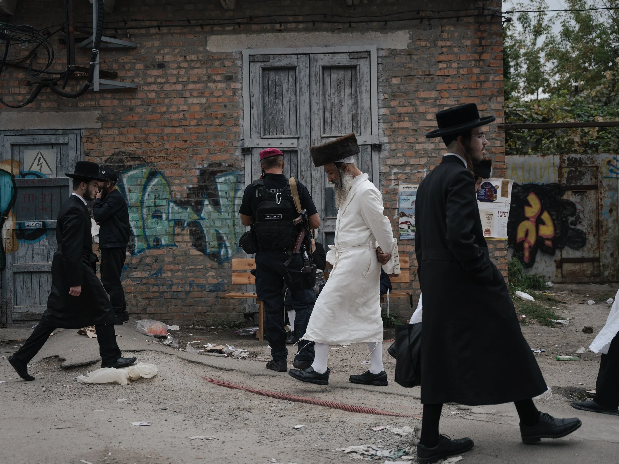 Group of Jewish men wearing traditional black hats and suits walking outside in front of a brick wall with graffiti. One man is dressed in white with a fur hat and long beard, walking among them.