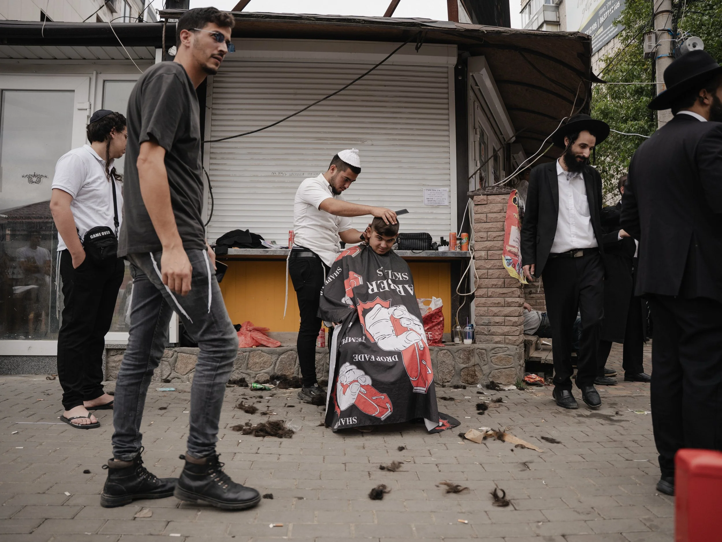 A street scene with several men, some dressed in black suits and traditional Jewish hats, standing and walking, with one man getting a haircut at a small outdoor barber stand. Hair clippings are scattered on the pavement.