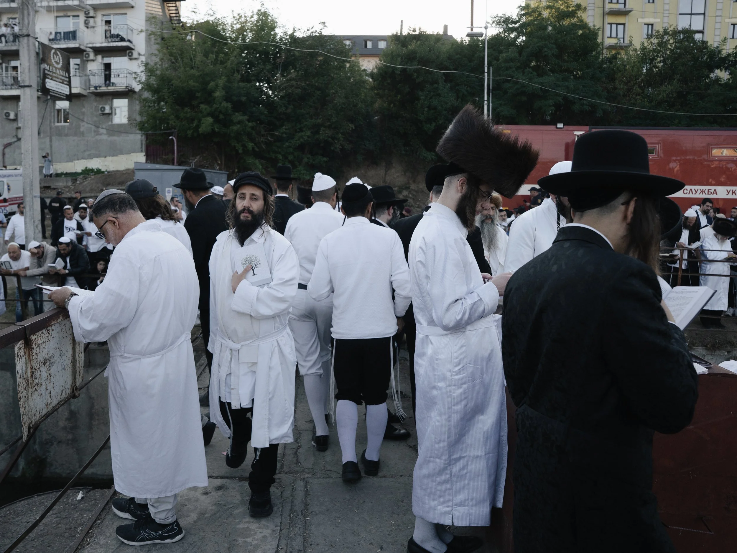 Group of men dressed in traditional Orthodox Jewish clothing, including black hats and white robes, gathered outdoors. Some men are reading from books, possibly prayer books.