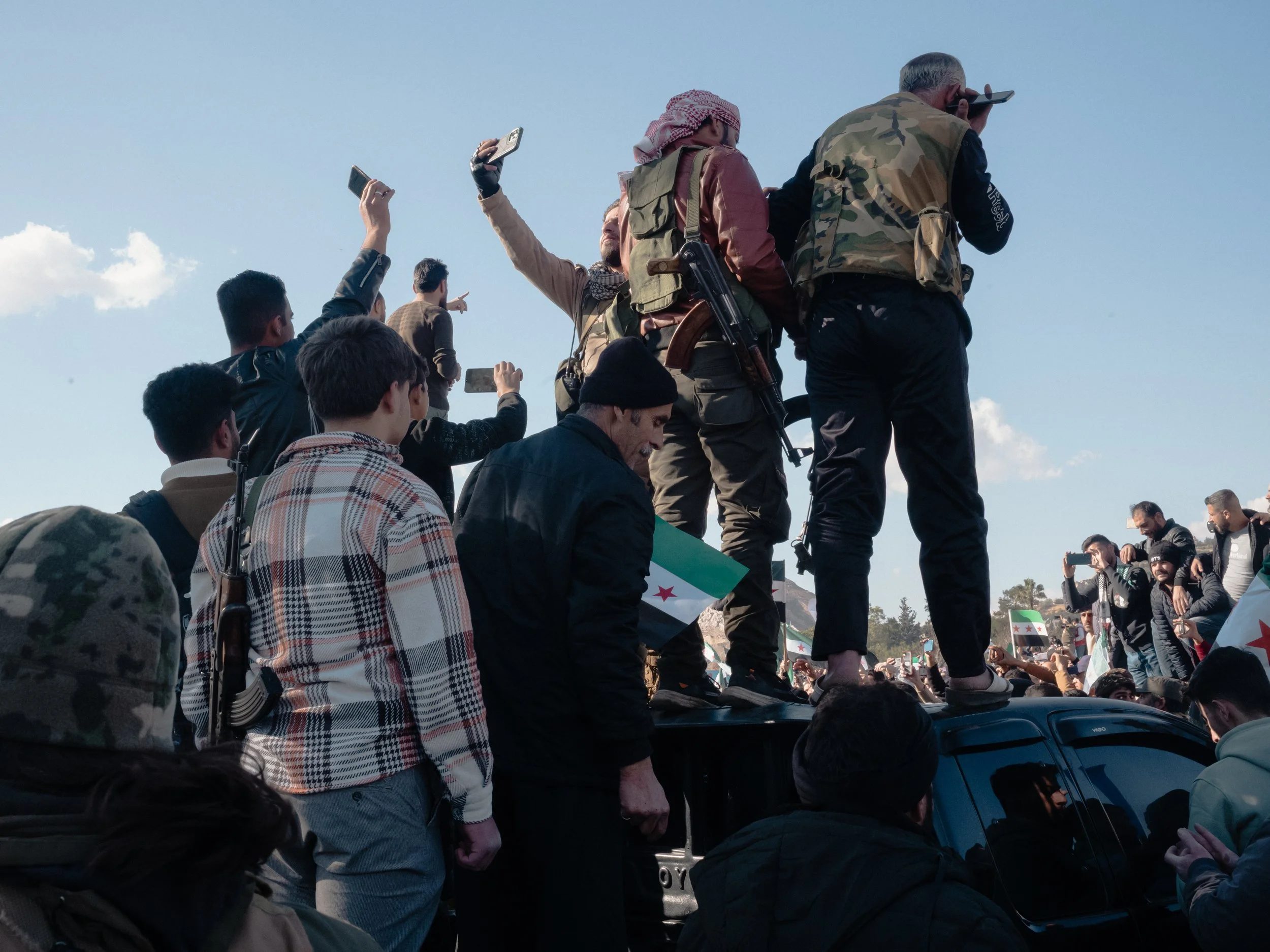 People gather around a procession with individuals standing on the roof of a vehicle, some holding smartphones, with a blue sky and scattered clouds in the background.