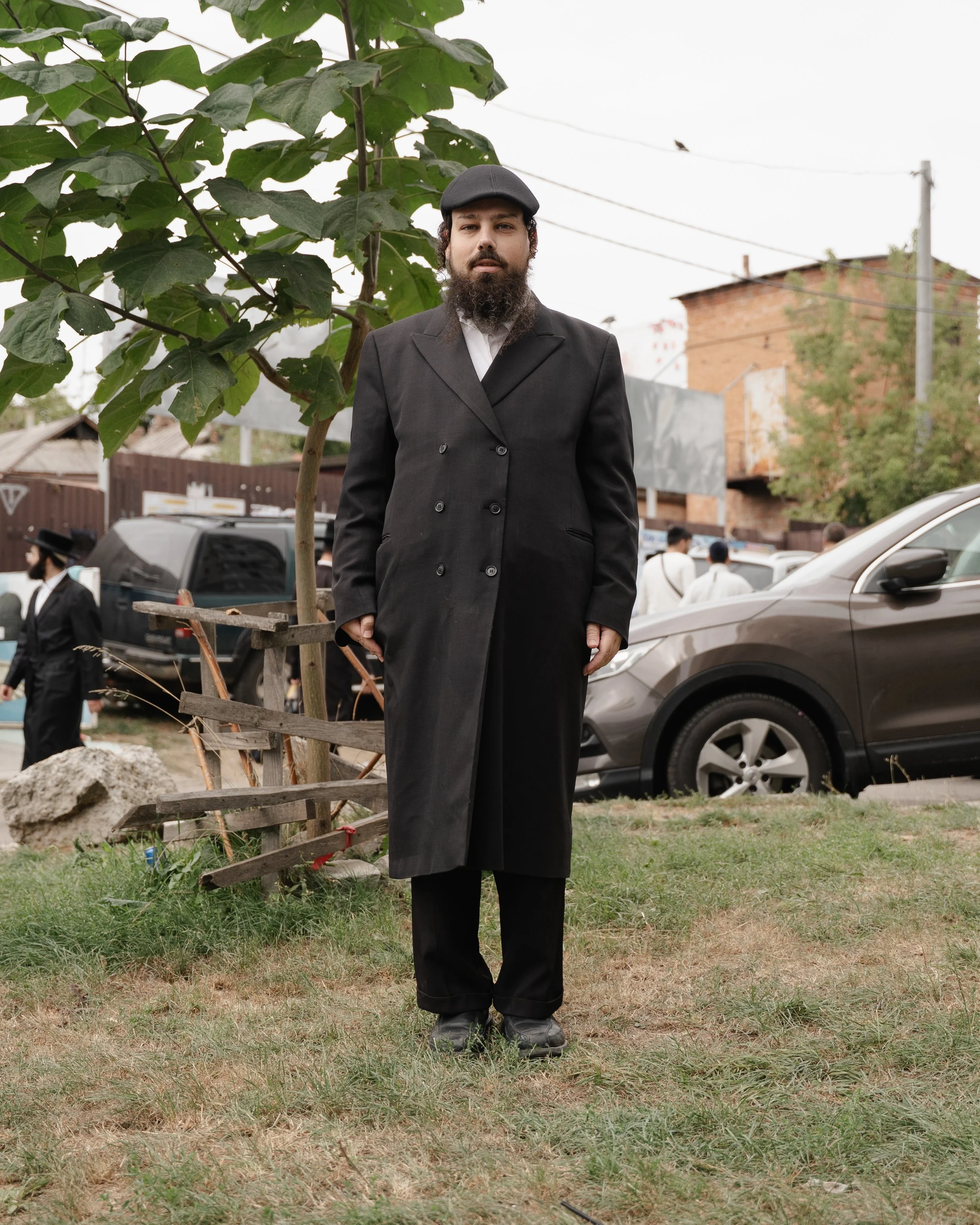 A man dressed in a black coat and black trousers with a gray flat cap, standing outdoors in a grassy area with trees and cars behind him. There are people in the background near parked cars, and a building with a brick wall and power lines are visible in the distance.